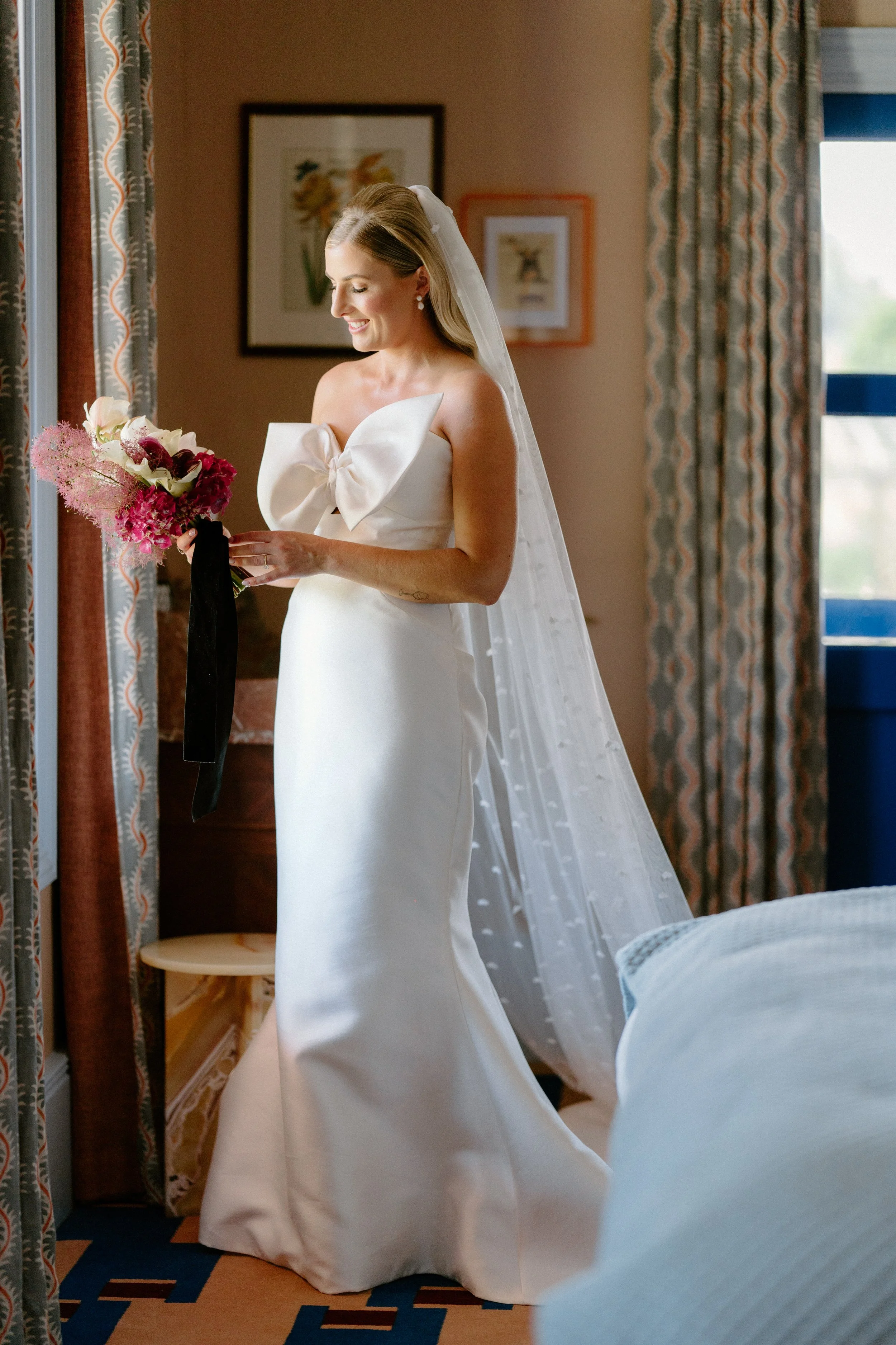 The Sienna, Hunter Valley Wedding Venue, Wedding Photos. A bride in a white wedding gown with a large bow on the front, holding a bouquet of flowers, standing in a room near a window with patterned curtains.