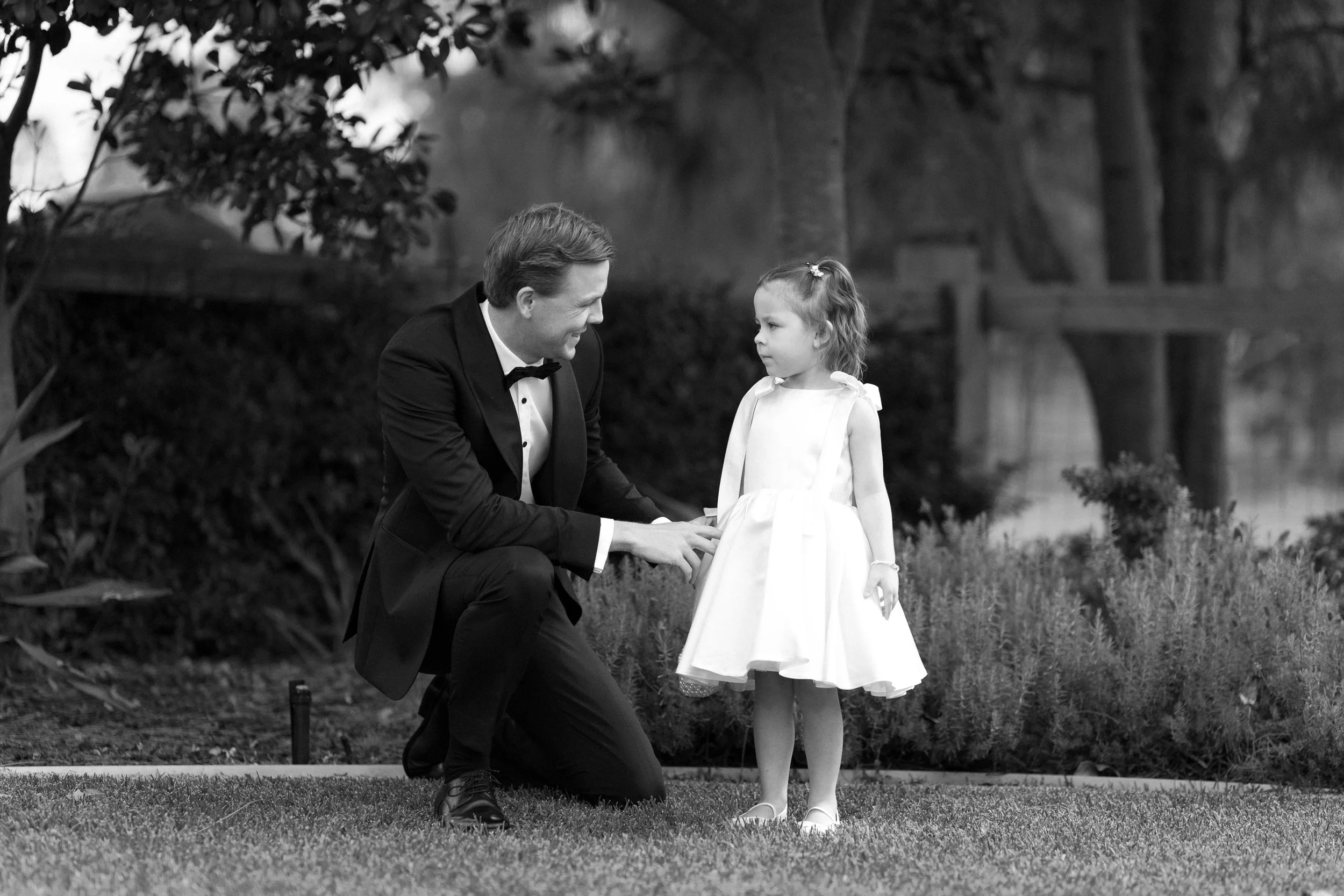 A man in a tuxedo kneeling down and holding the hands of a young girl in a white dress outside near a pond and trees.