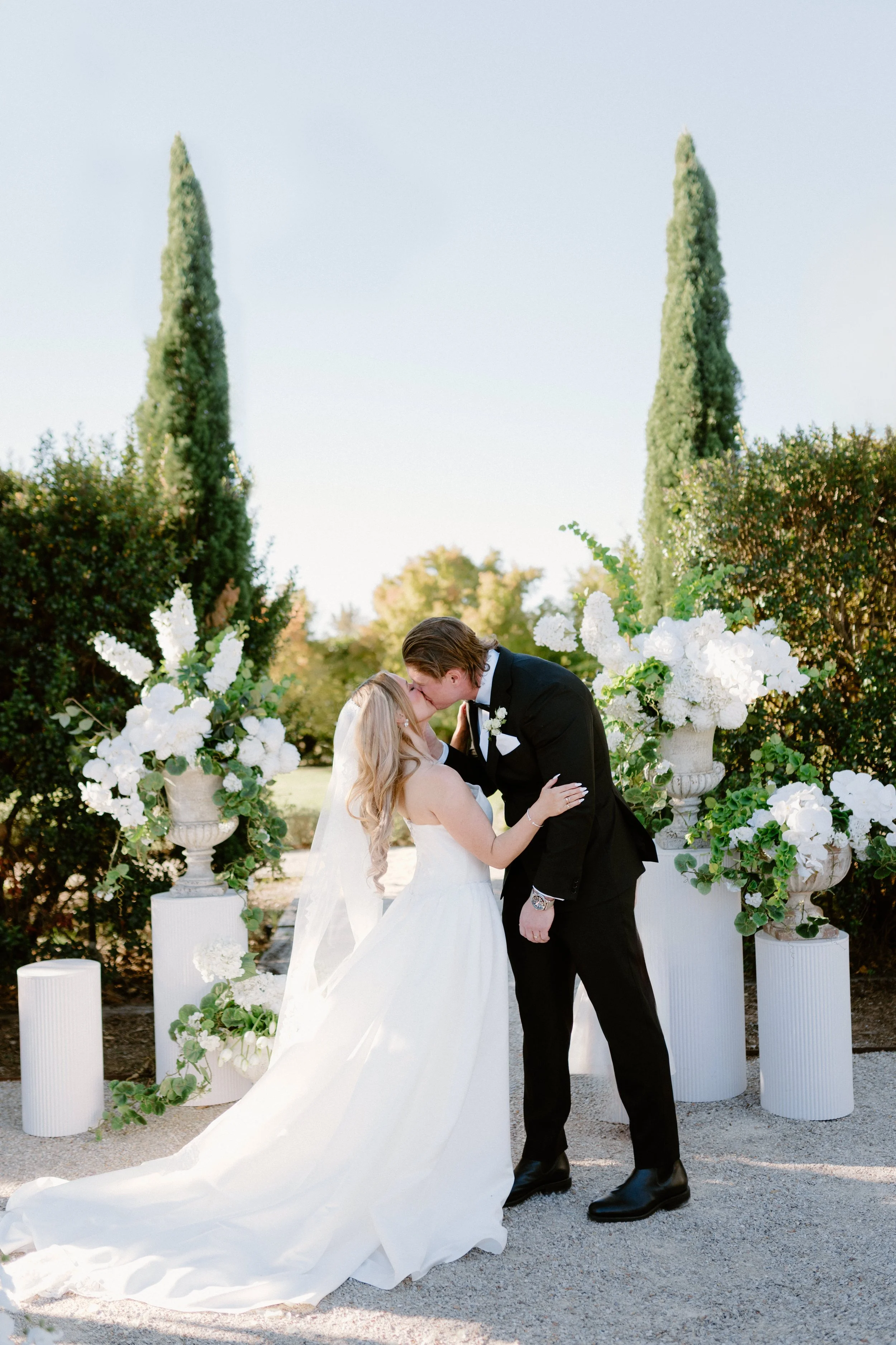 A bride and groom sharing a kiss during their outdoor wedding ceremony, surrounded by large white floral arrangements in urns, with tall cypress trees in the background.