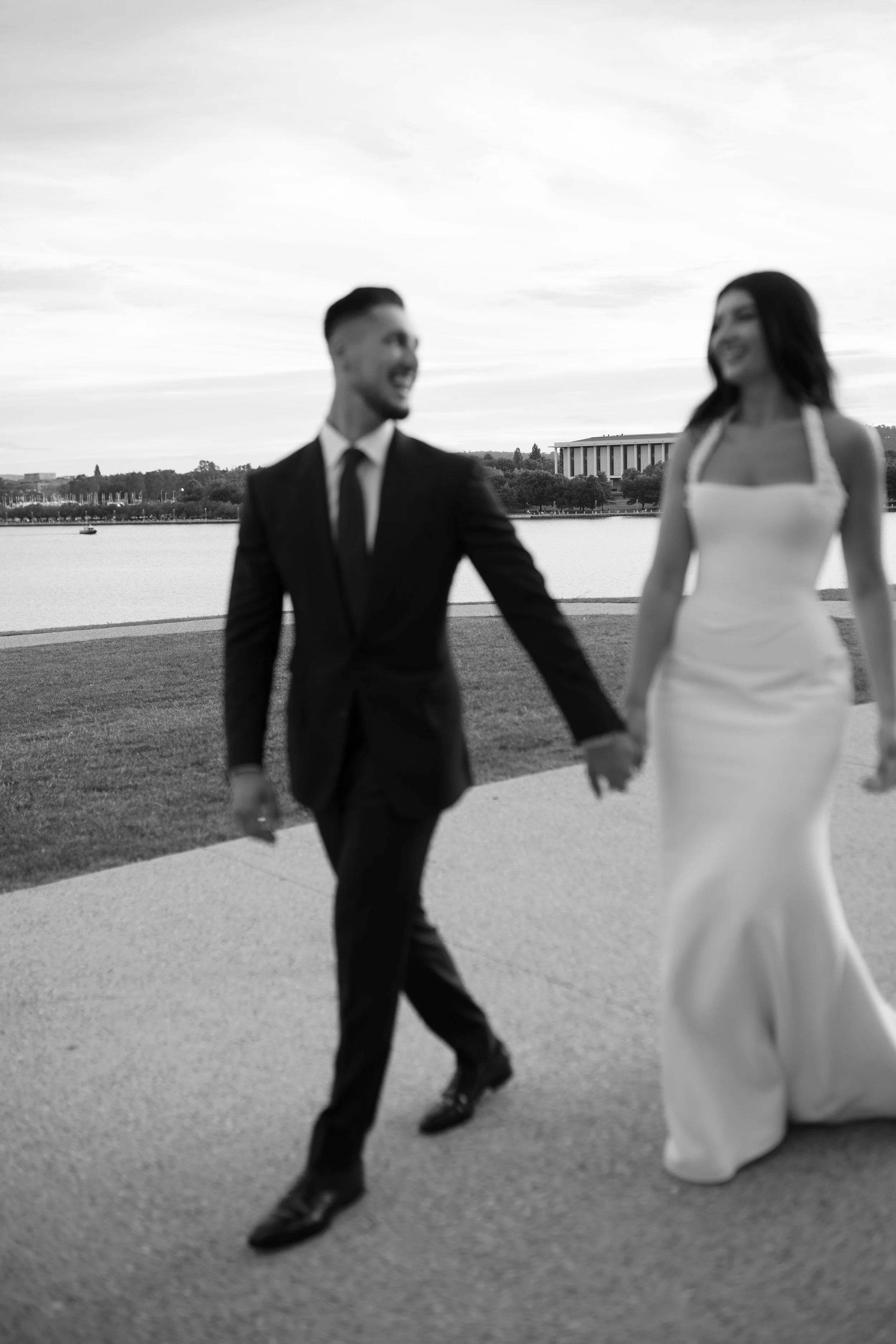 A couple dressed in wedding attire holding hands by a lake with a city skyline and a large building in the background, black and white photo.