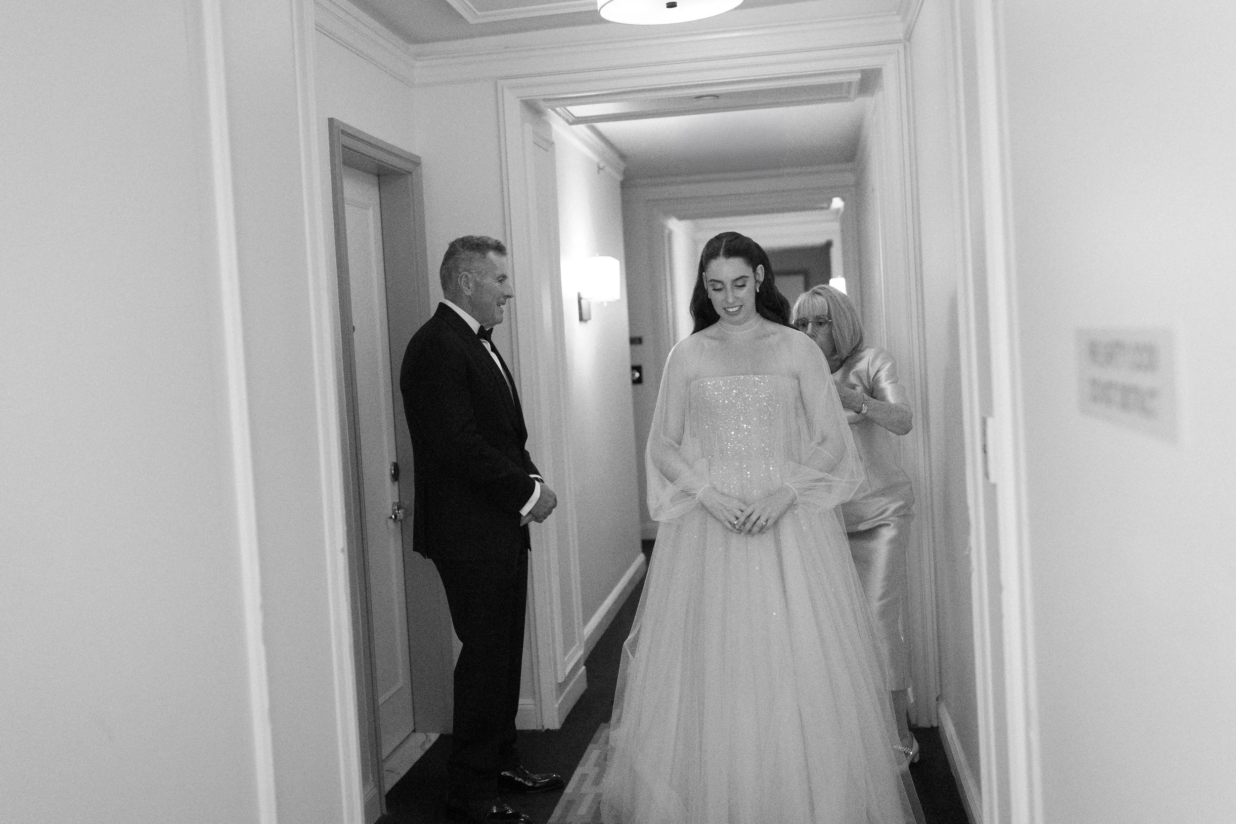A woman in a wedding dress is smiling in a hallway while two people, an older man in a tuxedo and an older woman in a satin dress, stand nearby, with the woman adjusting the dress behind her.