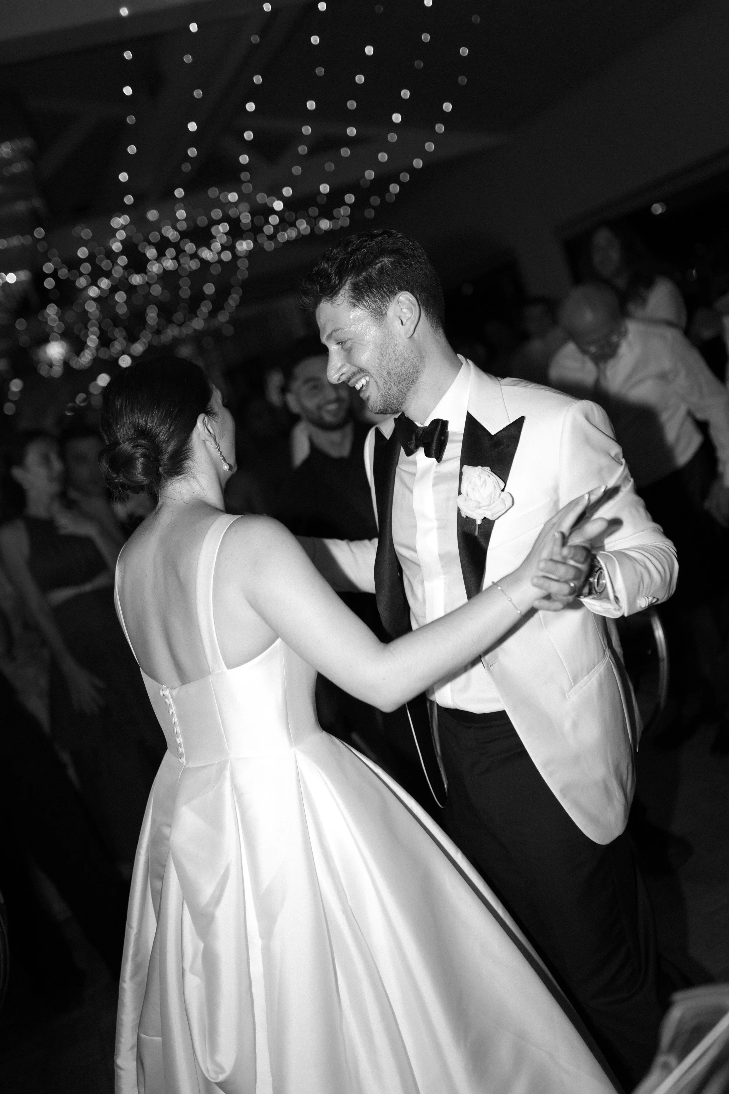 A black and white photo of a bride and groom dancing at their wedding reception, surrounded by guests, with string lights overhead.