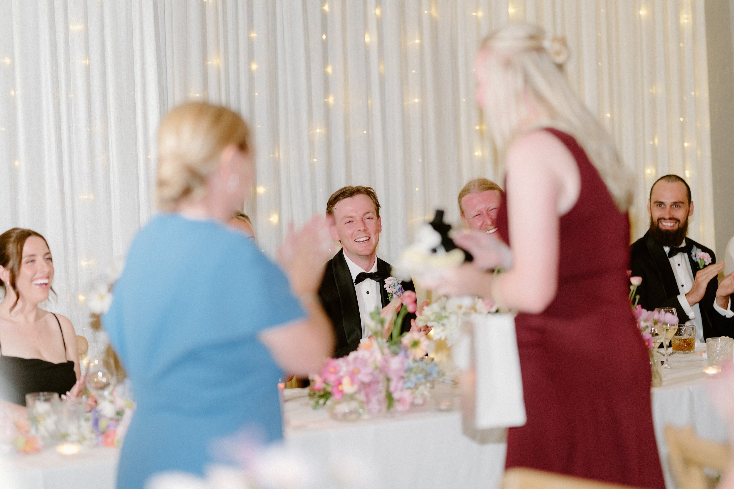 Guests at a wedding reception seated at a table decorated with pink and white flowers, with a woman giving a speech or toast. The background has white curtains with string lights.