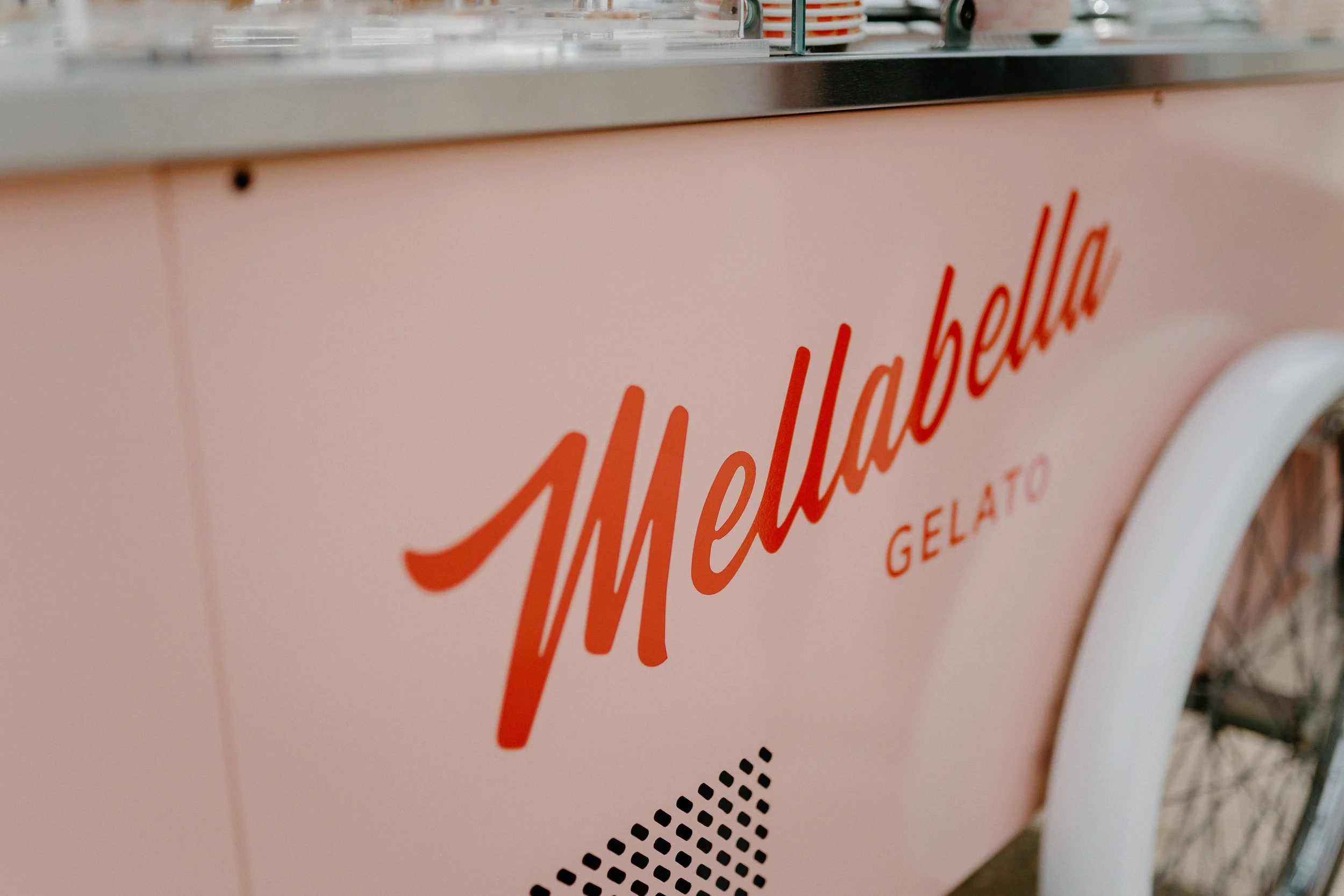 Close-up of an ice cream cart with the word 'Mellabella' written in red cursive, and 'Gelato' underneath. The cart is pink with a white wheel on the right side.