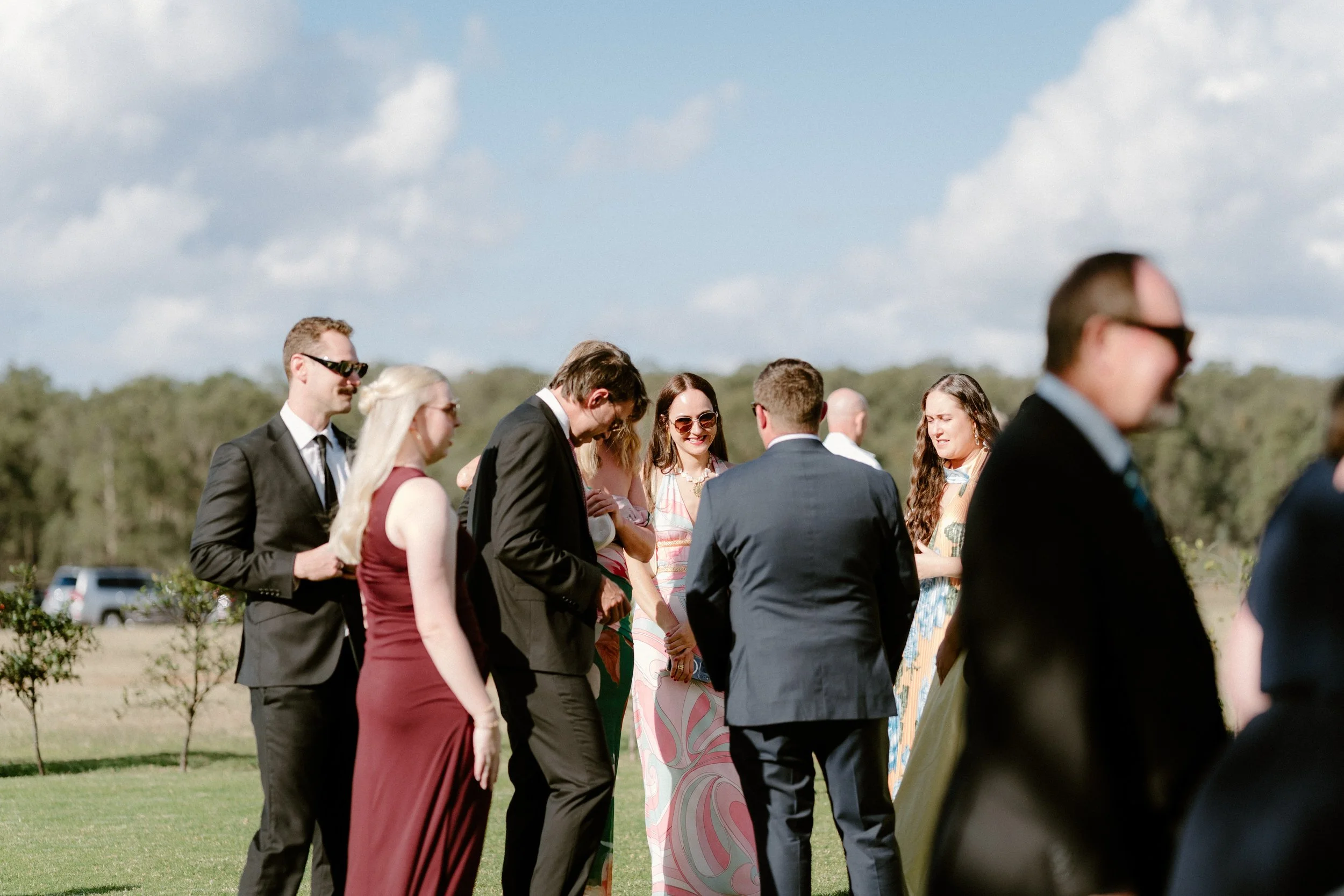 A group of well-dressed people standing outdoors in a grassy area under partly cloudy sky.