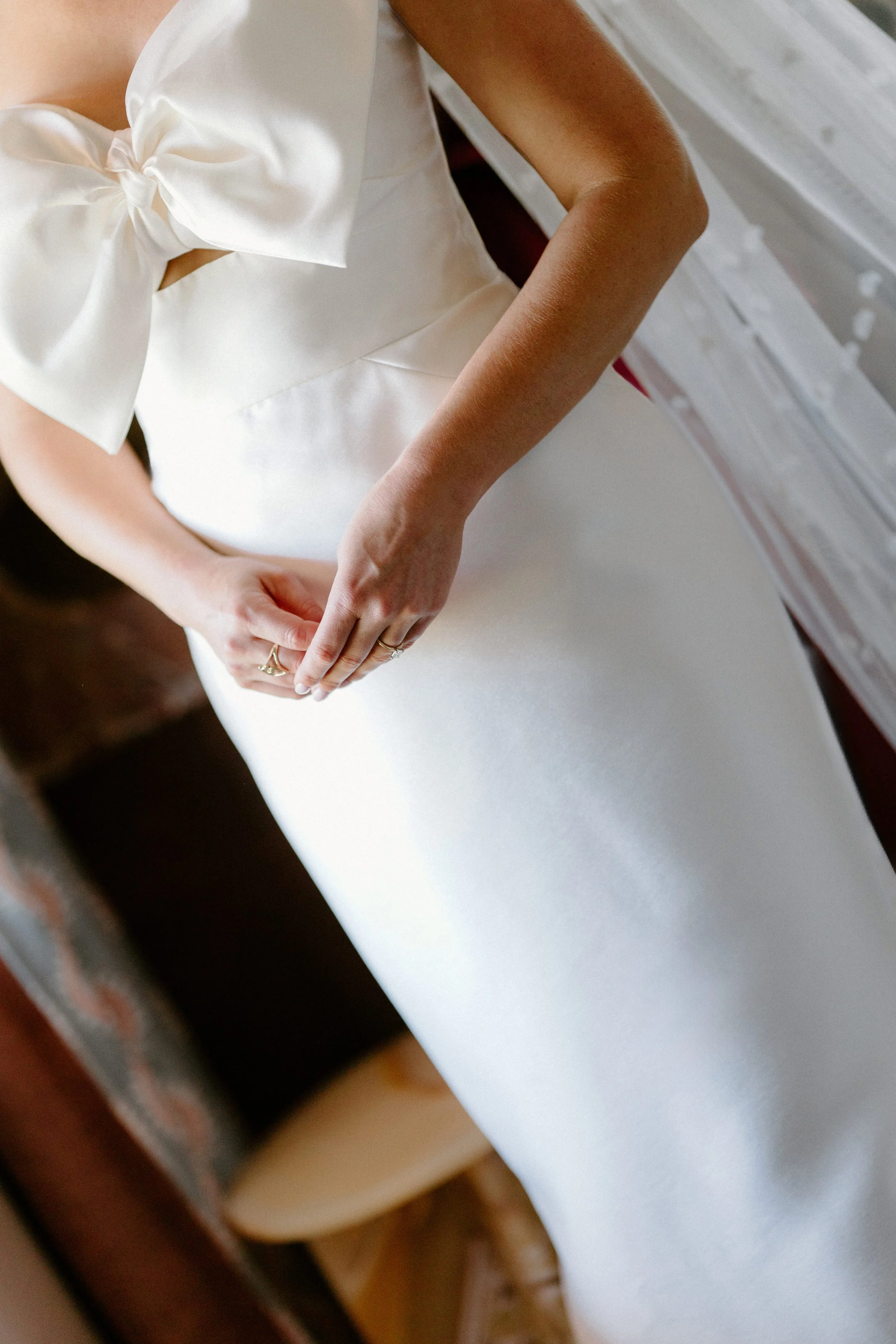 Close-up of a woman in a white satin dress with a bow, standing indoors. Her hands are clasped in front of her, and she is wearing rings.