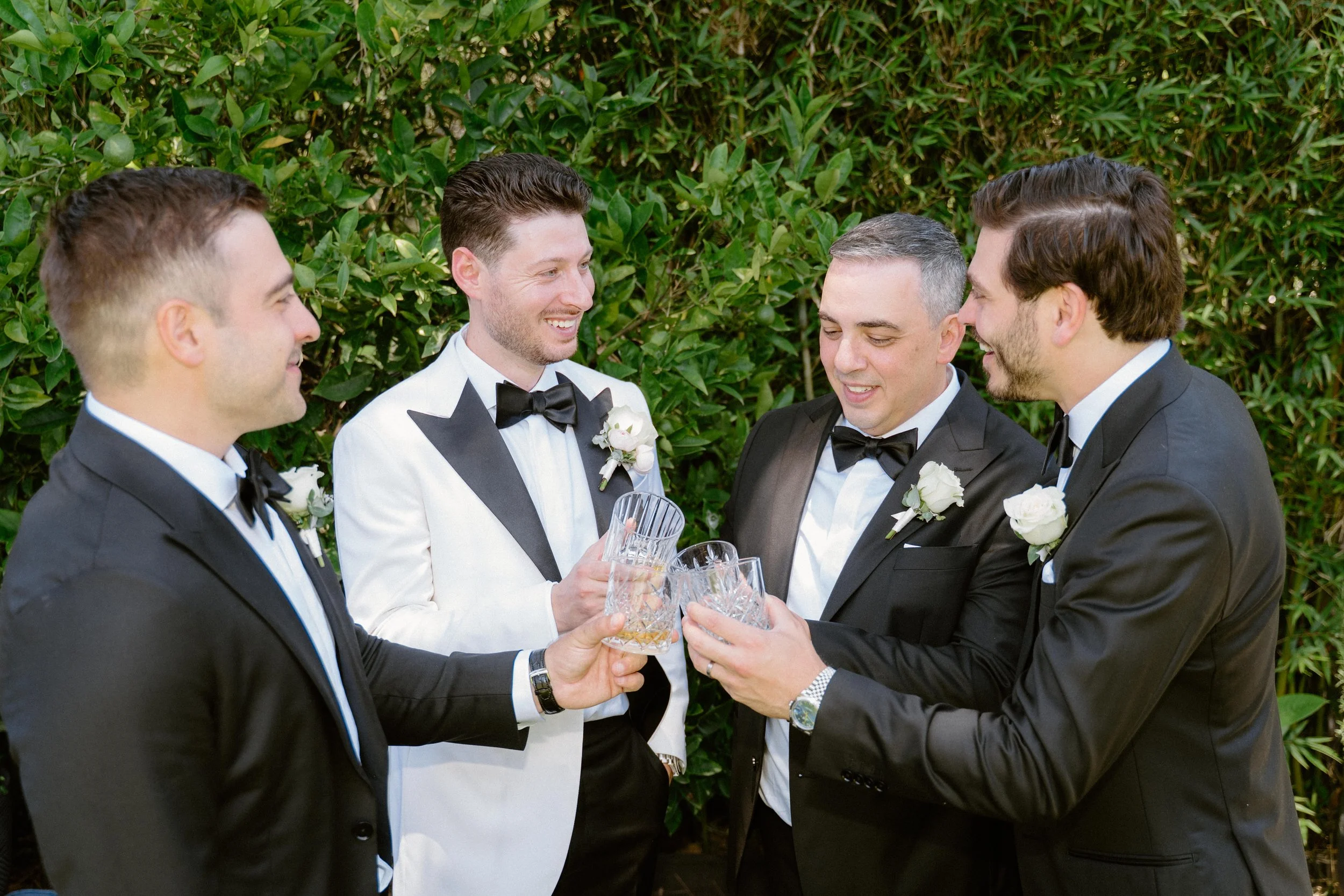 Five men in tuxedos with boutonnières, holding glasses of drinks, smiling and toasting together outdoors, with greenery in the background.