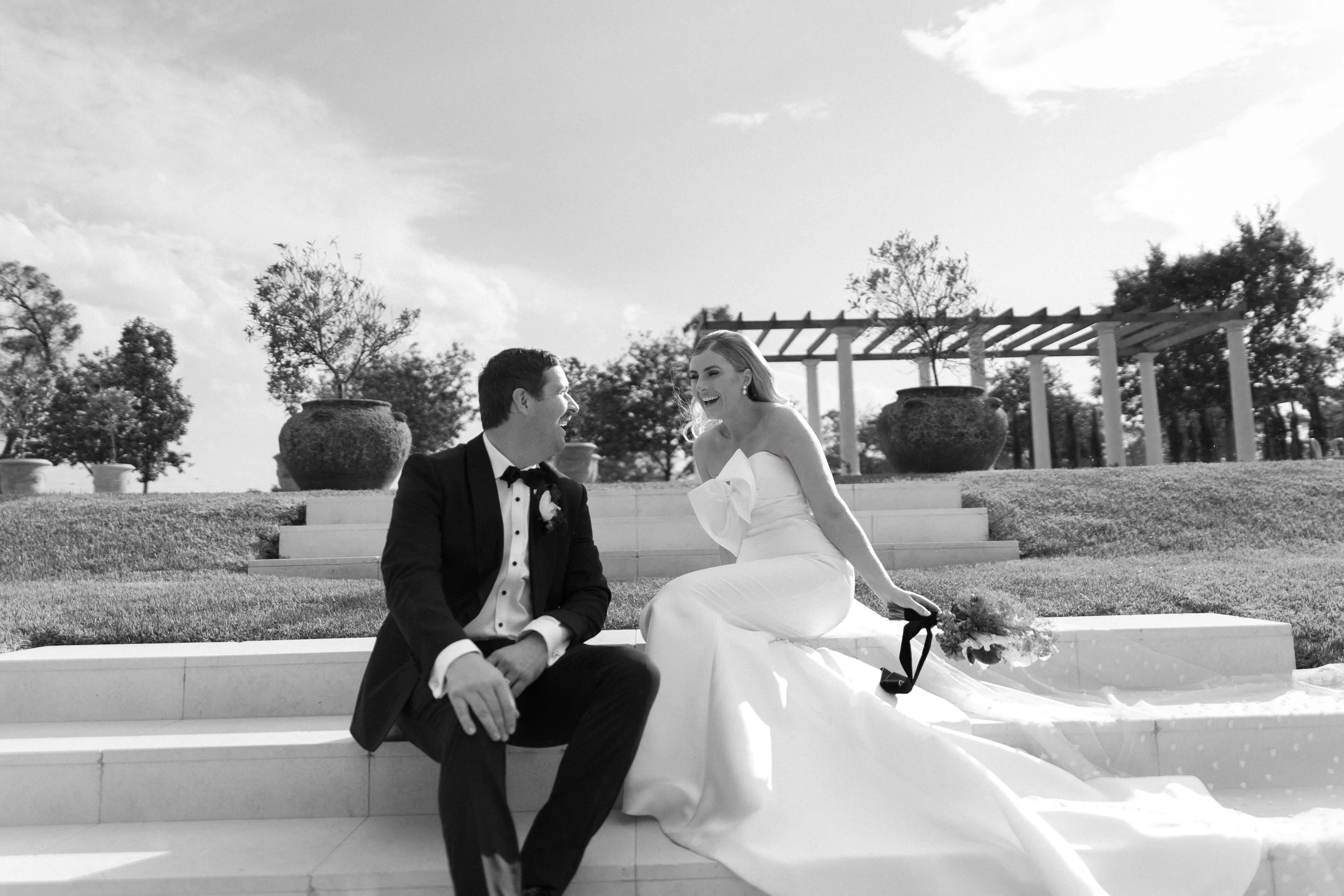 A black and white photo of a bride and groom sitting on steps outdoors, smiling at each other, with the bride holding a bouquet and wearing a strapless gown, and the groom in a tuxedo.
