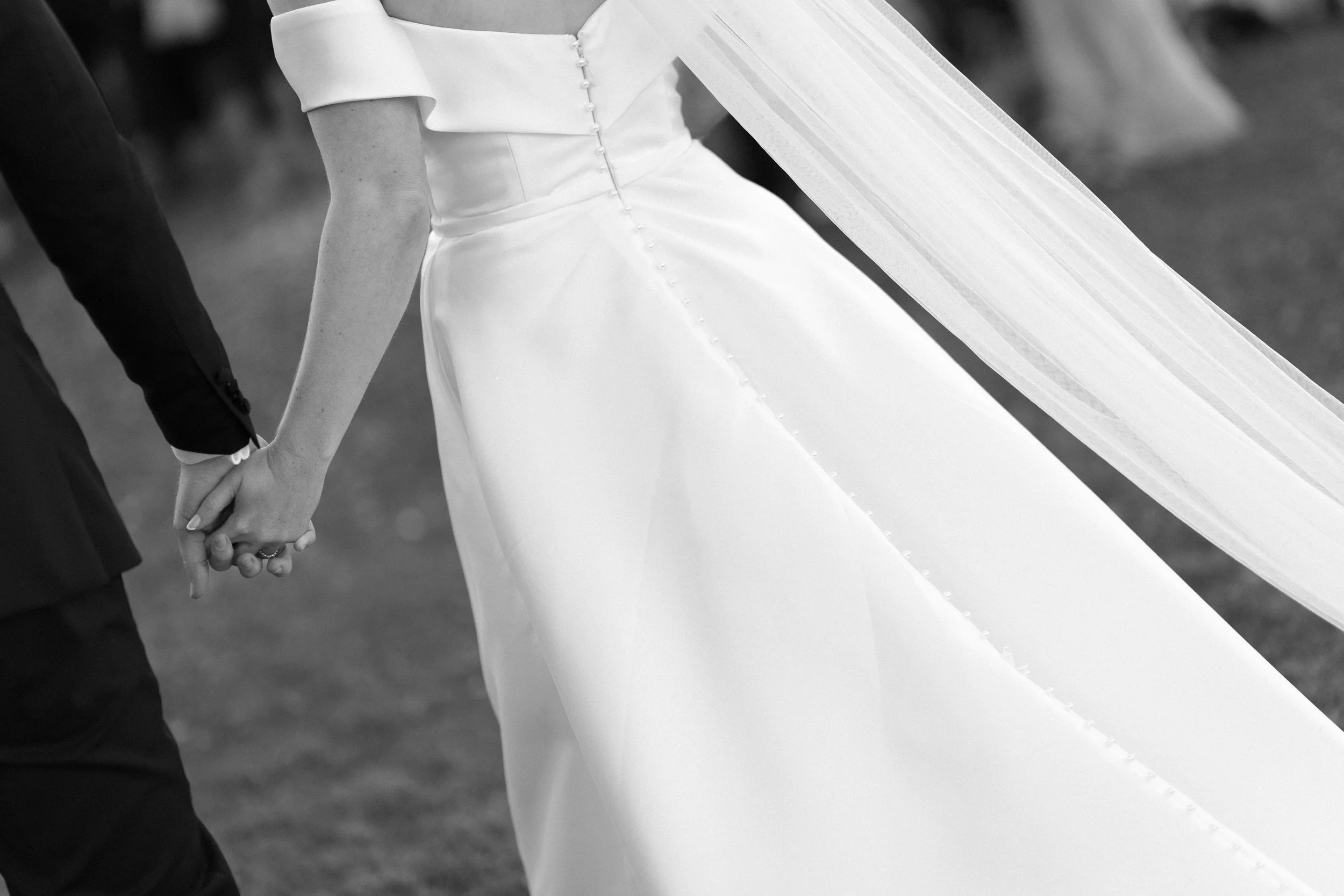 Close-up of a bride and groom holding hands during a wedding ceremony, with the bride wearing a white wedding gown and veil, and the groom dressed in a dark suit.