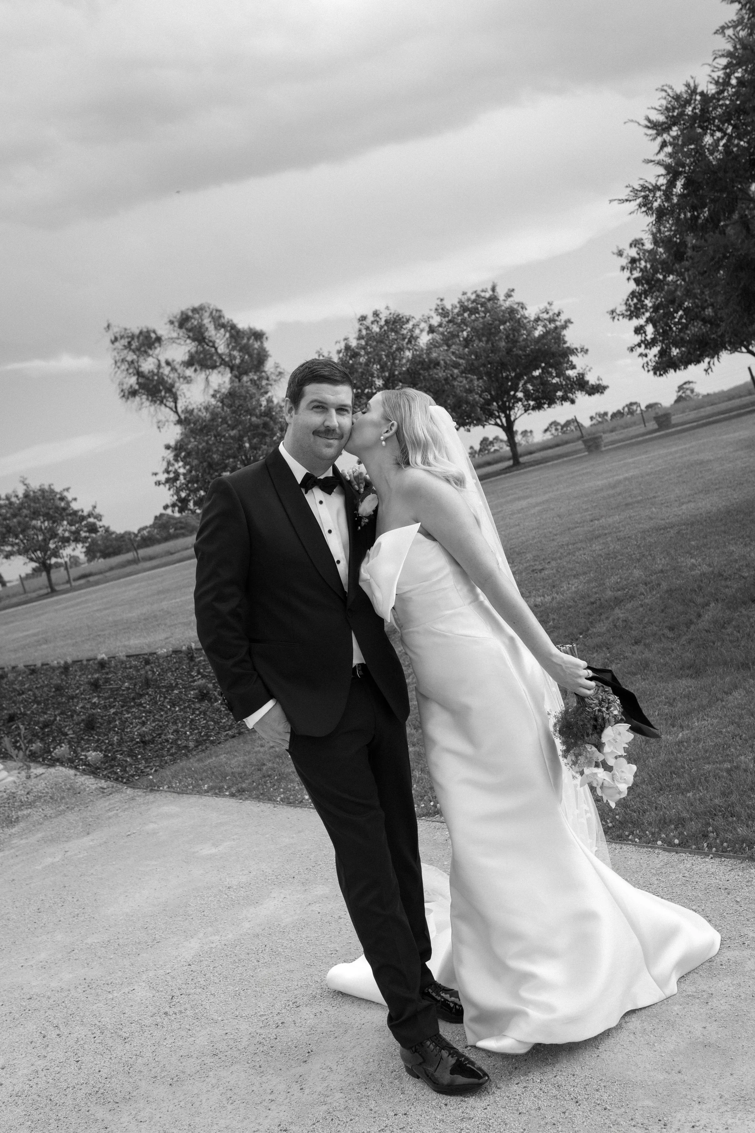 Black and white photo of a bride kissing a groom on the cheek outdoors on their wedding day. The groom is wearing a tuxedo and the bride is in a wedding gown holding a bouquet.