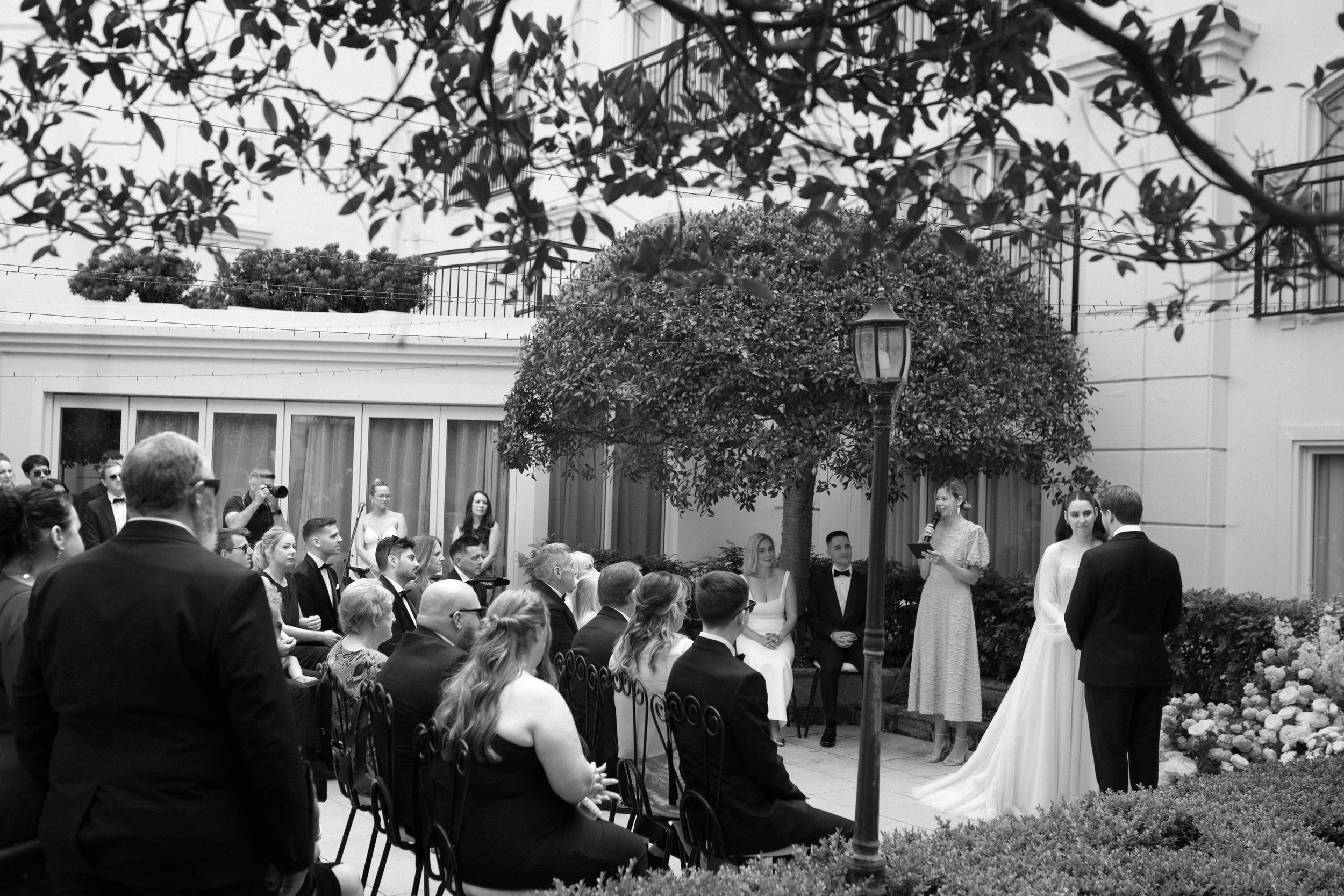 A wedding ceremony taking place outdoors in a garden or courtyard with guests seated and standing; a bride and groom are standing together, with a woman reading vows or giving a speech. There are trees, bushes, and a streetlamp in the scene, and the 