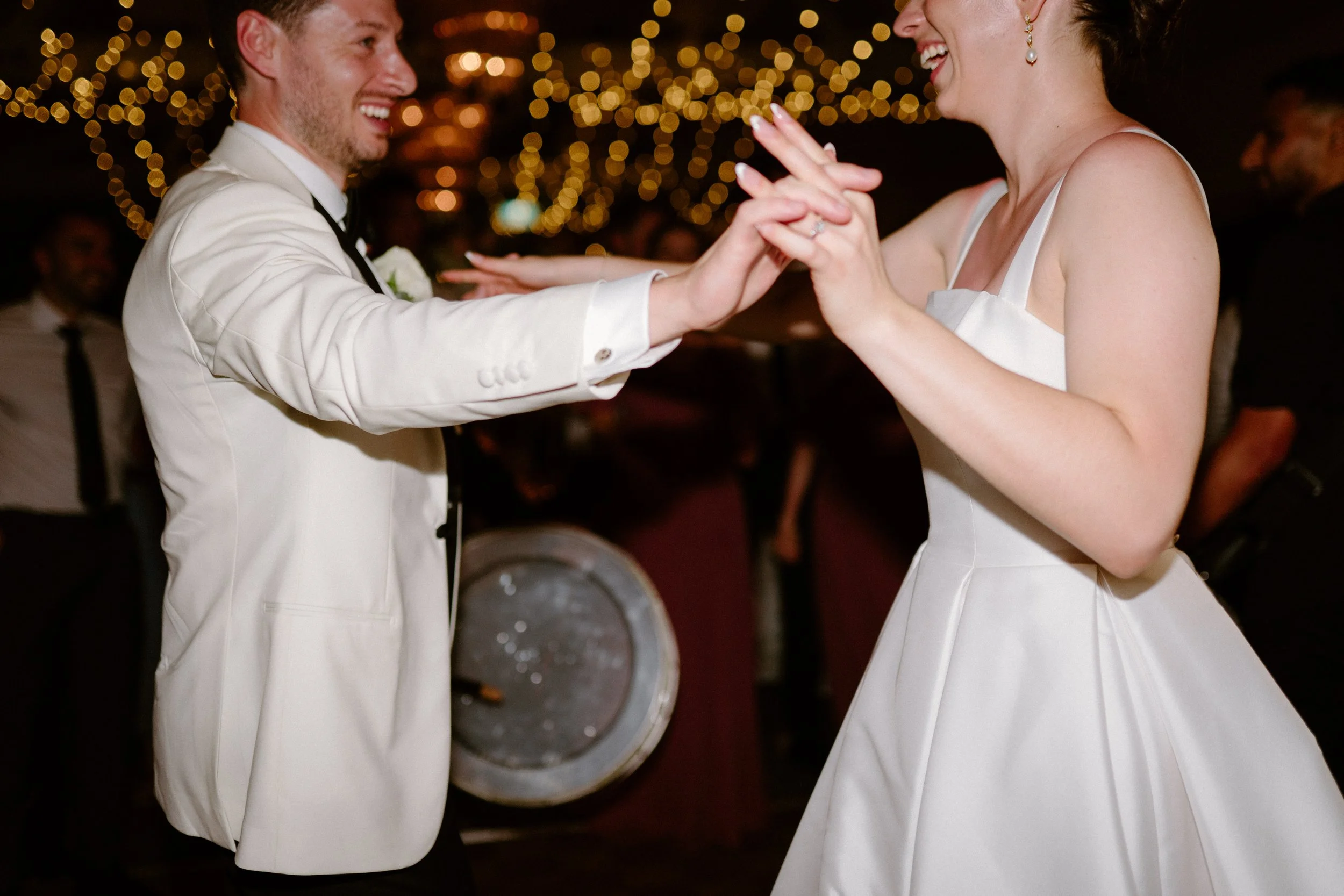 A bride and groom dancing at their wedding reception, smiling and holding hands, with string lights in the background.