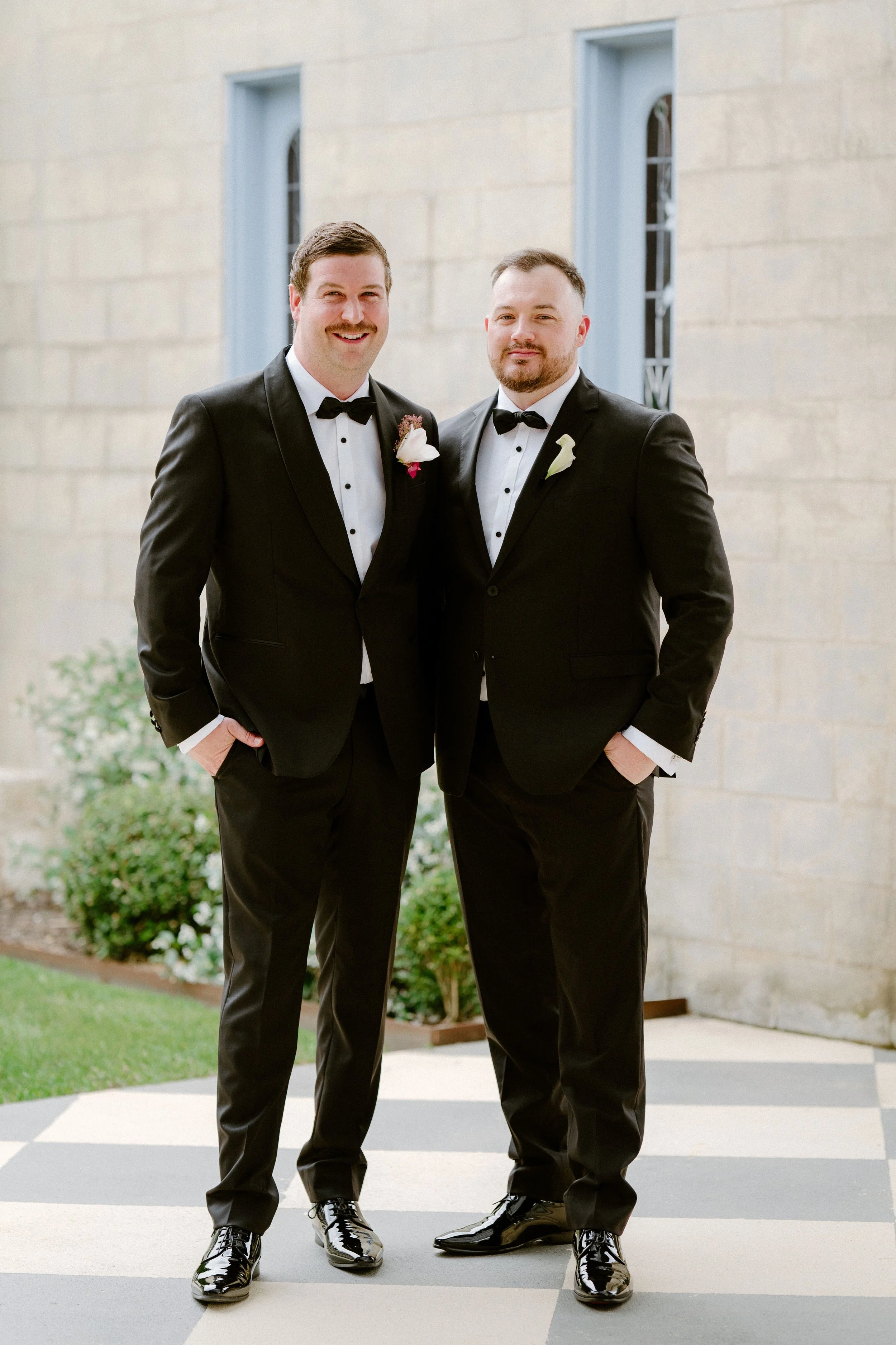 Two men in black tuxedos with bow ties and boutonnières stands side by side outdoors in front of a building with light-colored stone walls and blue-framed windows.