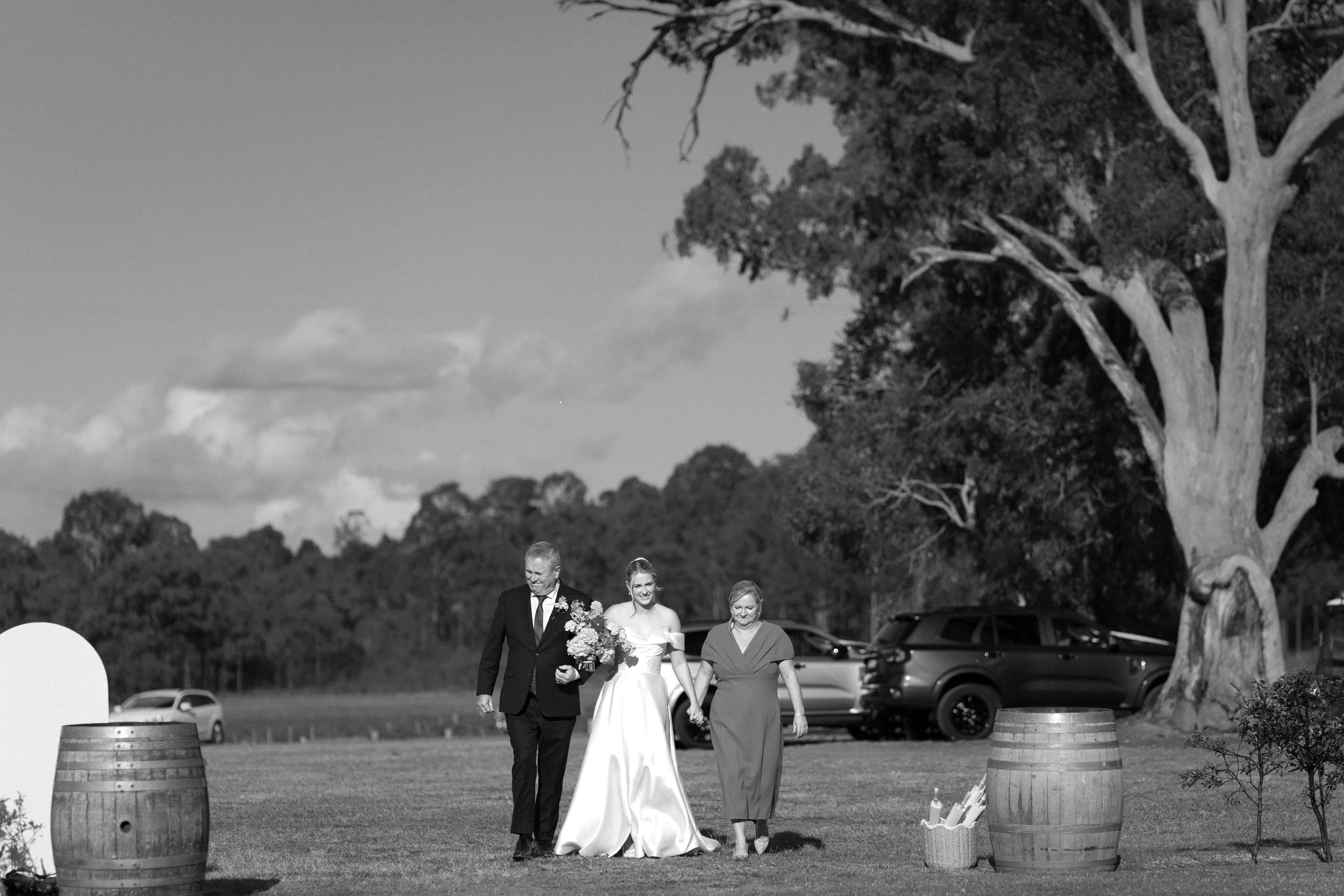 A black-and-white photo of a wedding ceremony outdoors. The bride and groom are walking together, surrounded by trees and parked cars, with two women walking alongside them, one older and one younger.