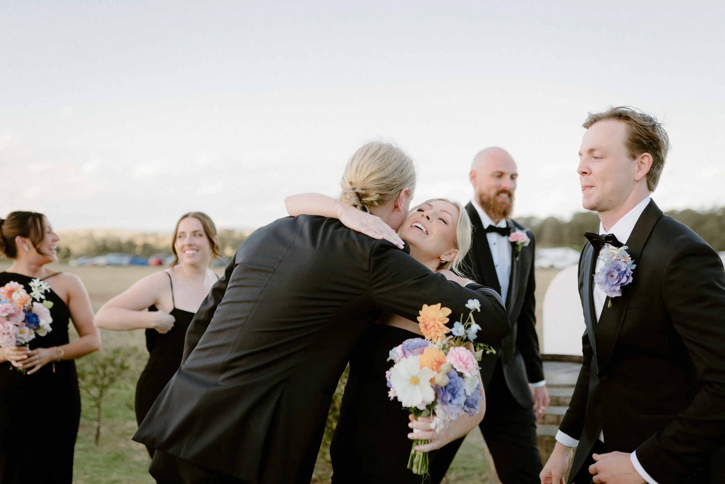 People at a wedding outdoors, including a woman in a black dress holding a floral bouquet, hugging a man in a tuxedo, all smiling.