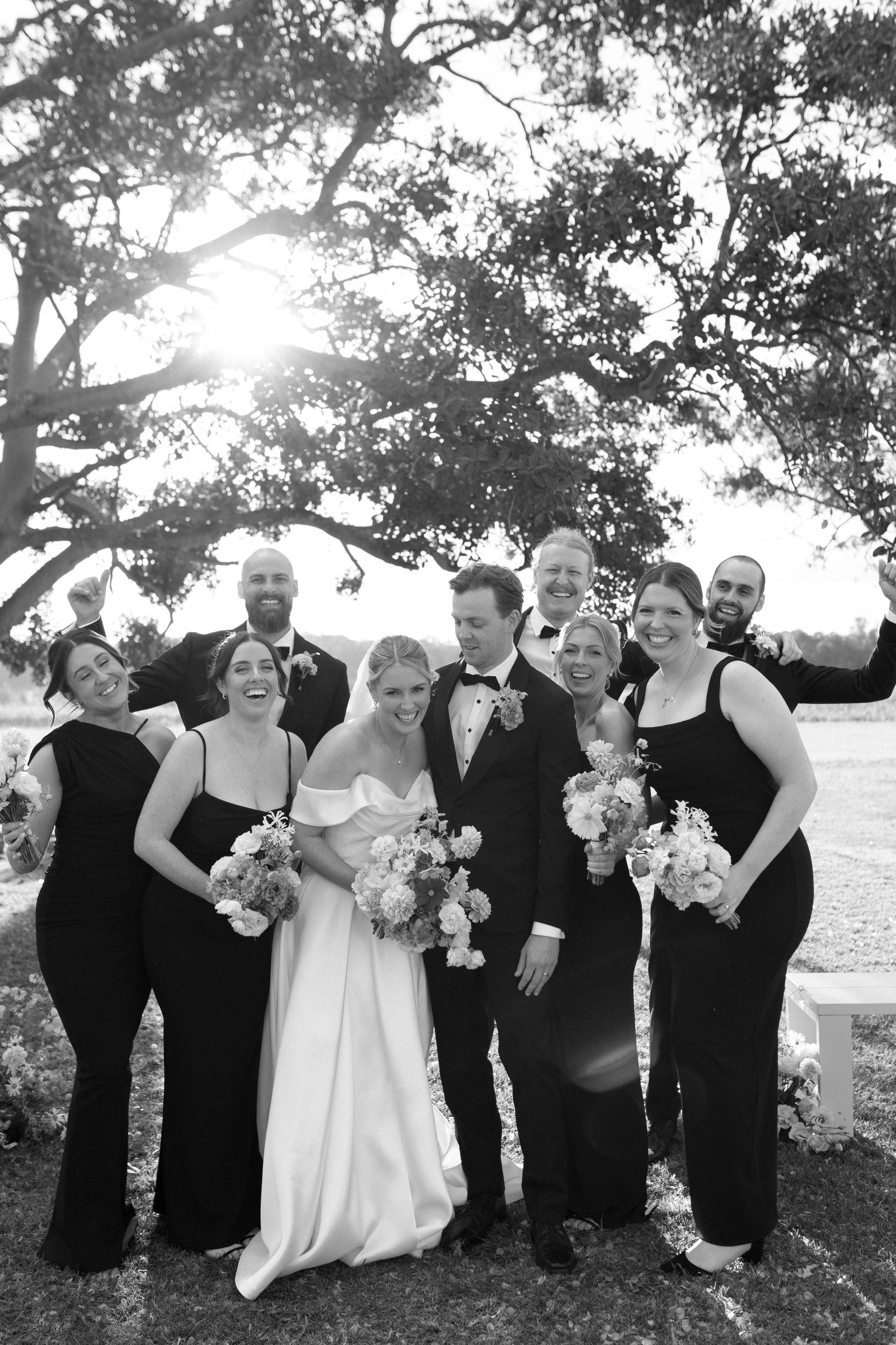A group of people dressed in formal attire standing outdoors, smiling, with trees and sunlight in the background, likely a wedding celebration.