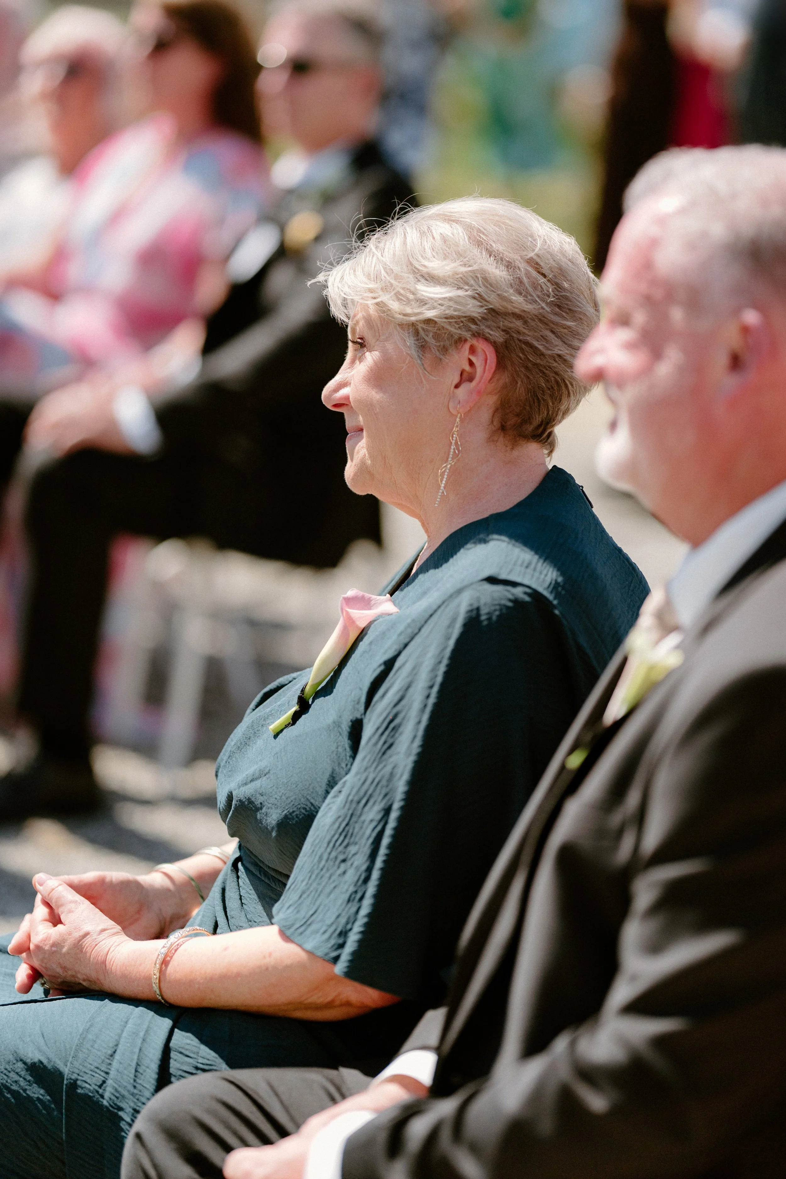 Profile of an older woman with short gray hair, wearing a dark dress and dangling earrings, sitting outdoors at a formal event, with a boutonniere on her dress and holding her hands in her lap. Other attendees are blurred in the background.
