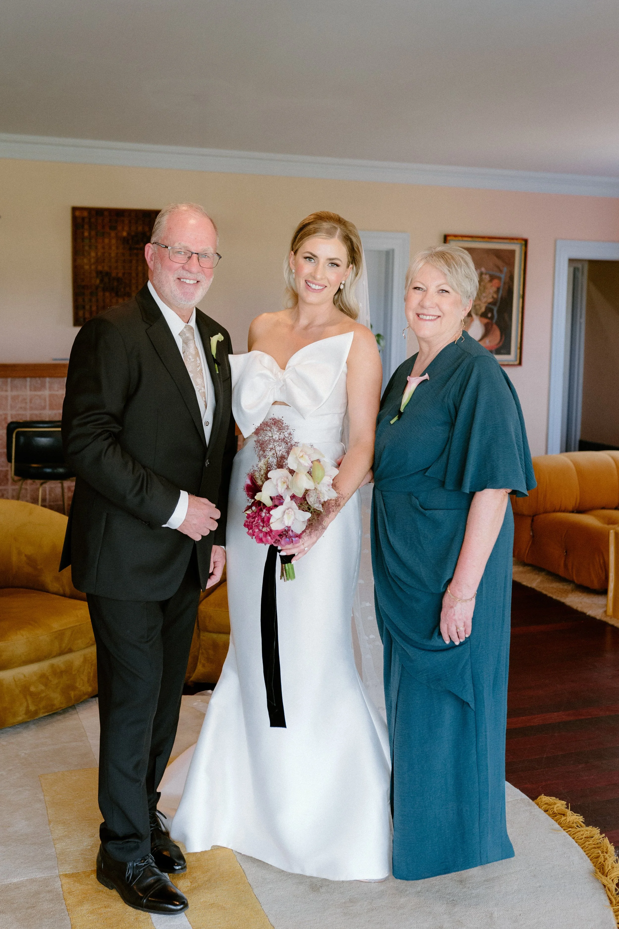 A bride in a white wedding dress holding a bouquet, standing between her father dressed in a black suit and her mother dressed in a teal dress, inside a living room.