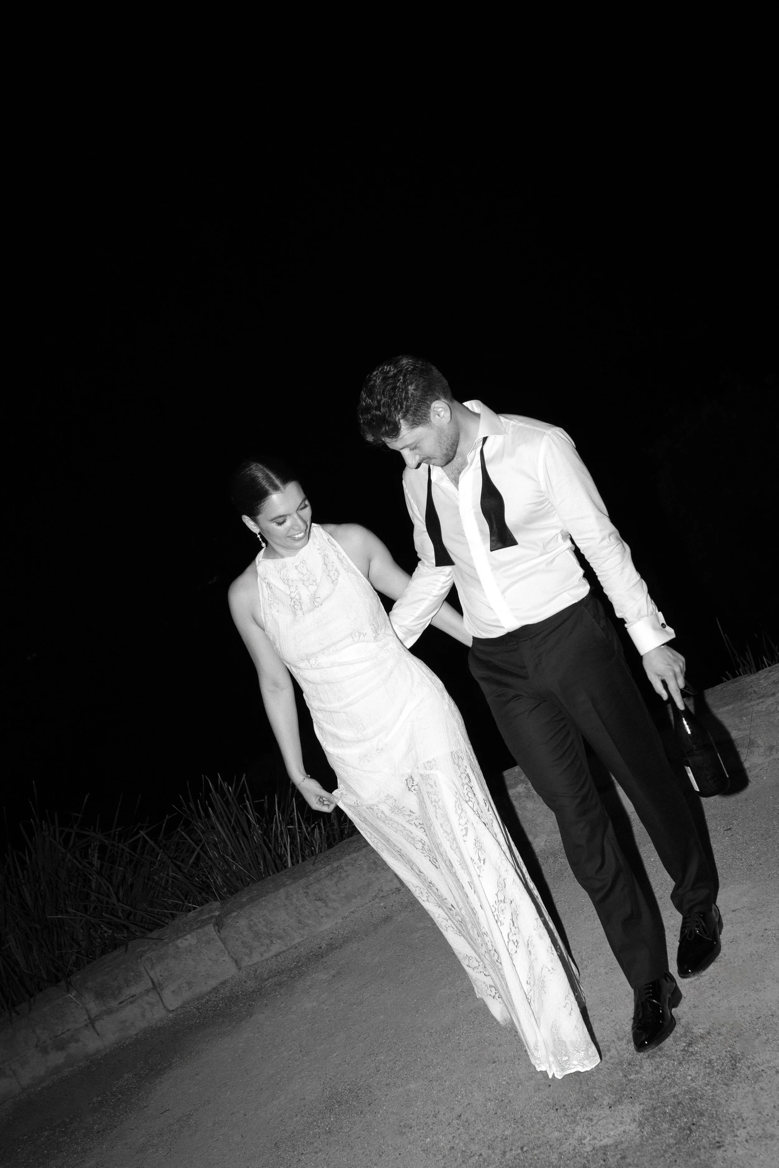 Sergeants Mess, Mosman Wedding Venue. Sergeants Mess Wedding Photos. Black and white photo of a man and woman walking together at night. The woman is wearing a long, elegant lace dress, and the man is dressed in a white shirt with a loosened tie, hol
