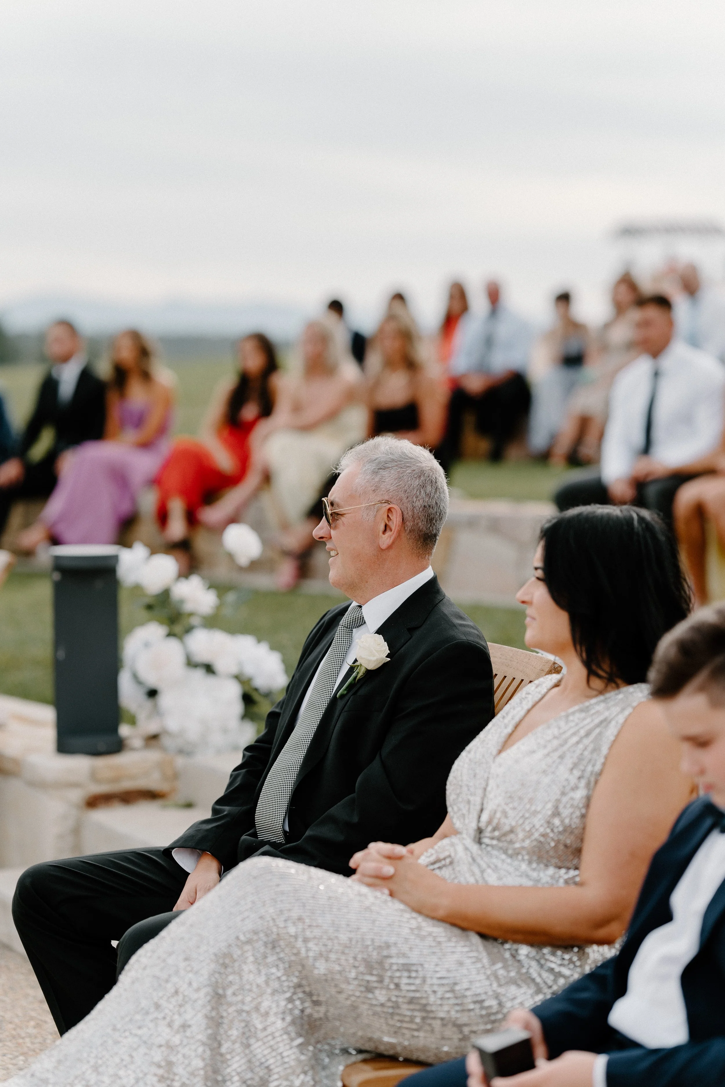 A wedding ceremony outdoors with guests seated on benches and a groom and bride sitting in the front row, smiling. The groom is wearing a black suit with a white boutonniere, and the bride is wearing a shimmering silver dress.