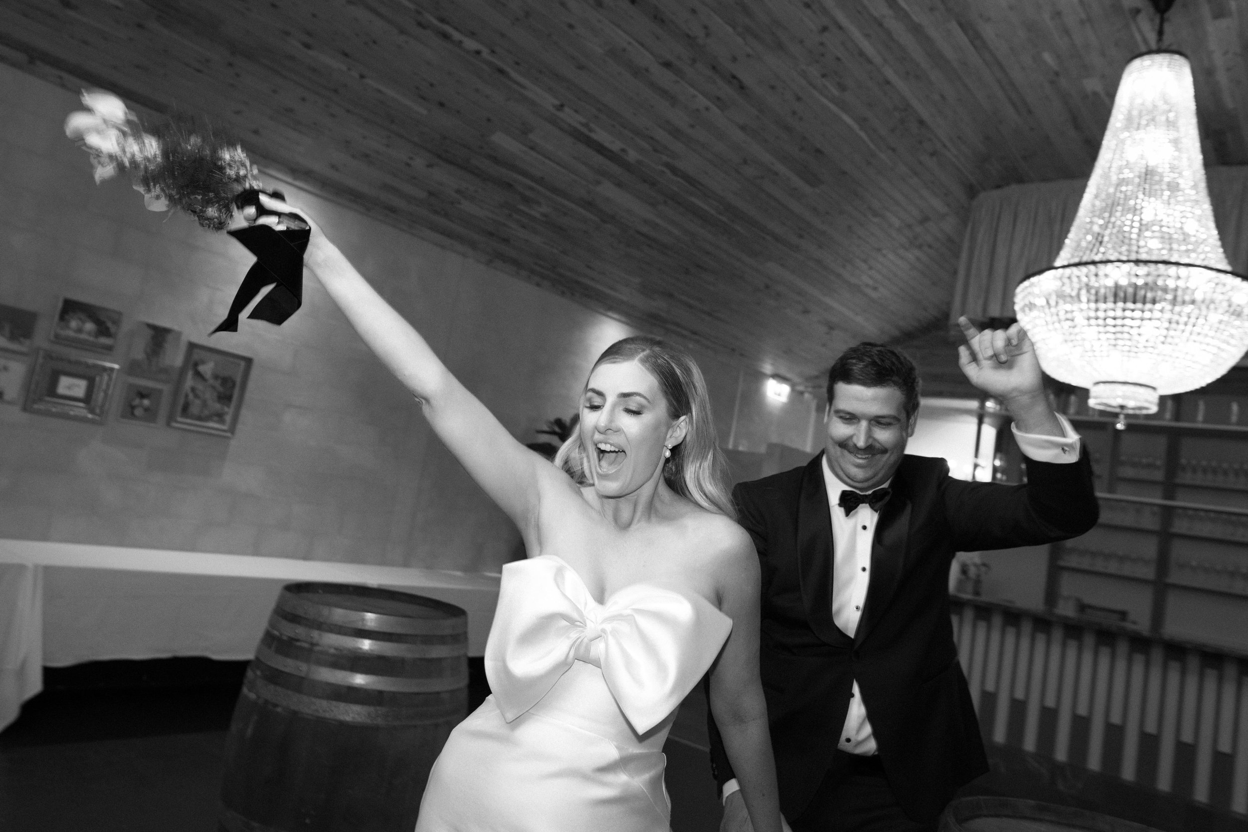 Black and white photo of a bride and groom celebrating at their wedding reception, with the bride waving a bouquet and the groom smiling.