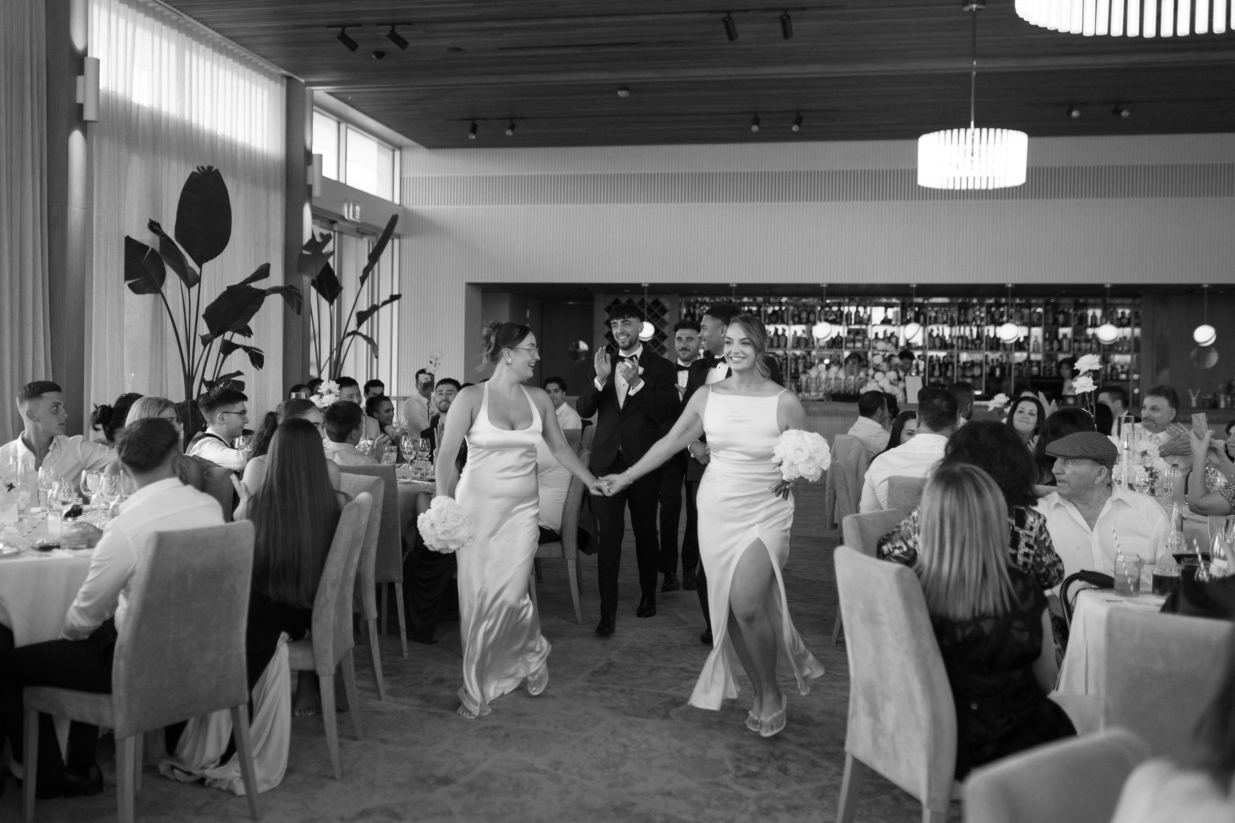 Black and white photo of a wedding reception with two brides holding hands and walking, surrounded by seated guests at decorated tables, with a bar in the background.