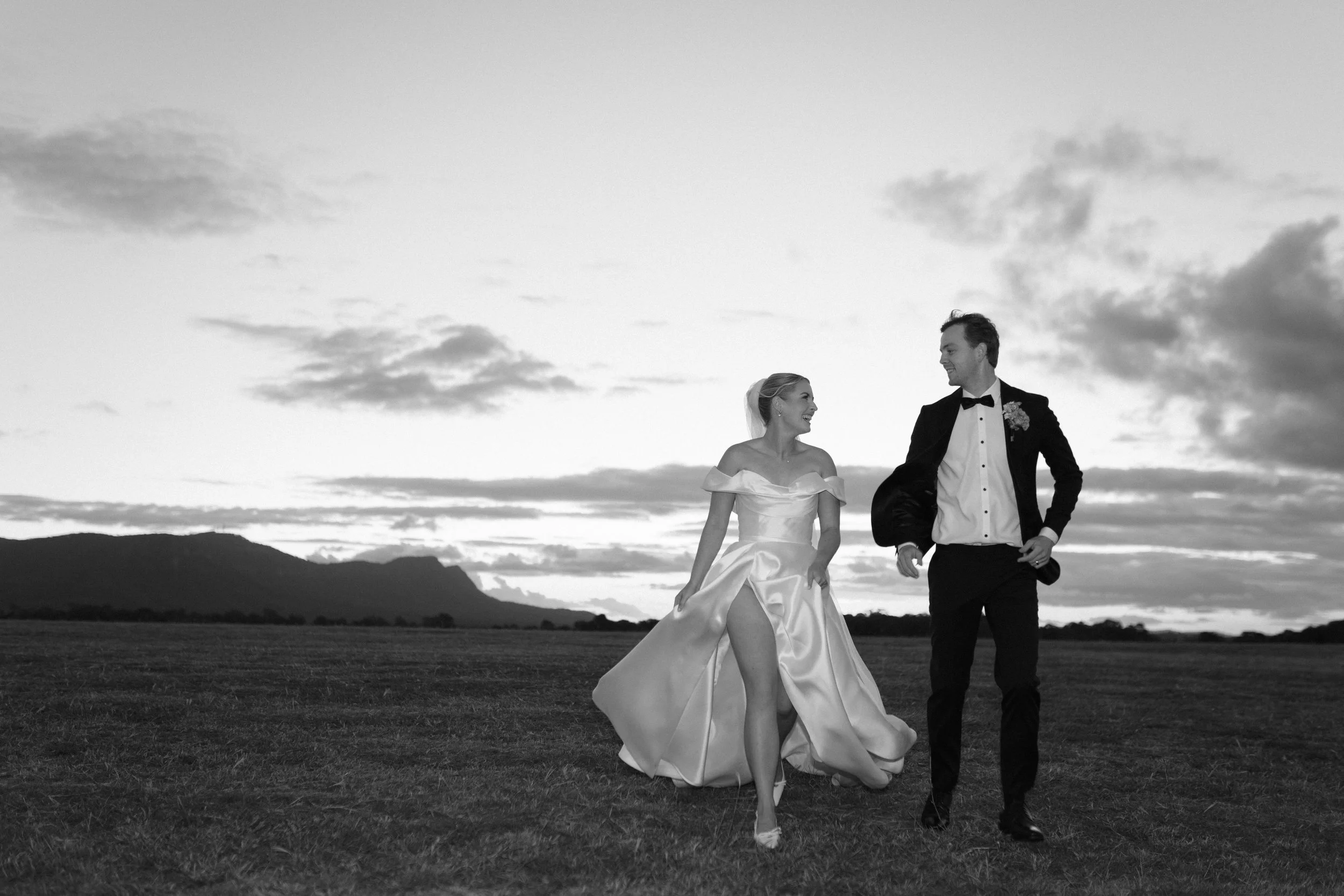 A bride and groom walking together outdoors during sunset, smiling at each other. The bride is in a strapless wedding gown, lifting her skirt slightly, and the groom is in a tuxedo with a bow tie, holding his jacket over his shoulder. The background 