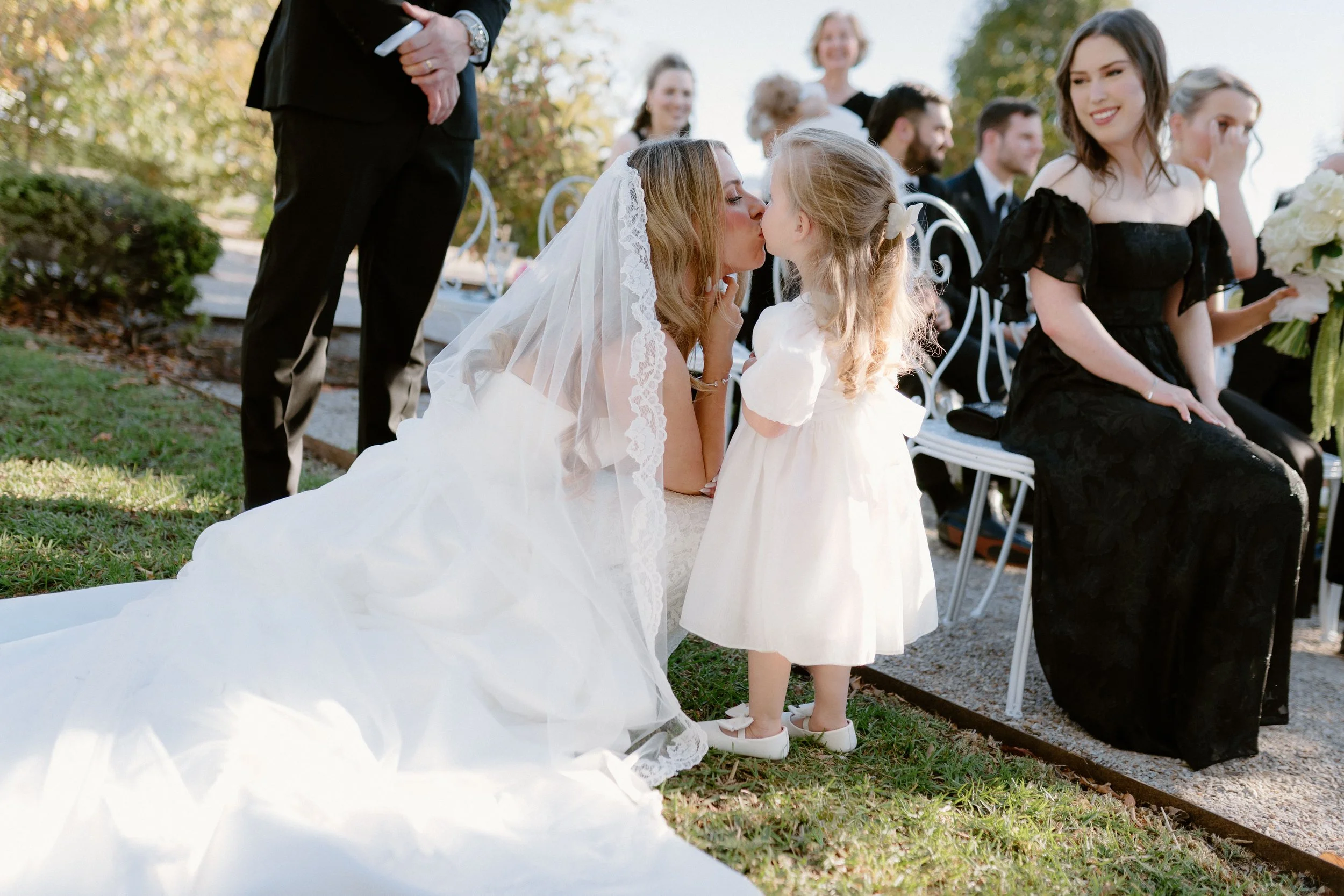 A bride in a white wedding dress and veil is kneeling and kissing a young girl in a white dress at an outdoor wedding ceremony. People are sitting in the background, dressed formally, with trees and sunlight.