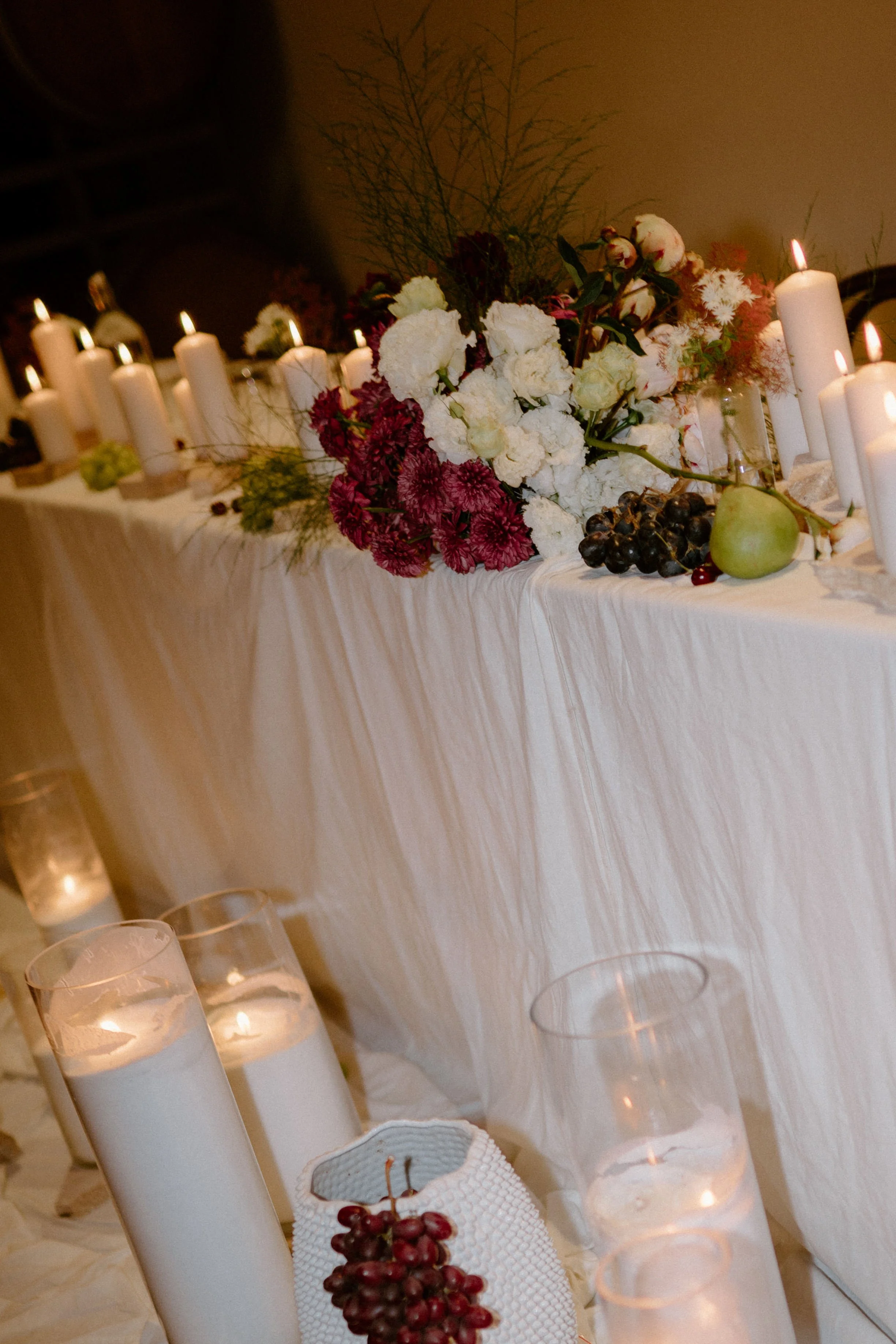 A table decorated with white candles, white and deep red flowers, grapes, apples, and other fruits, set on a tablecloth with a backdrop of a beige wall and some decorative branches.
