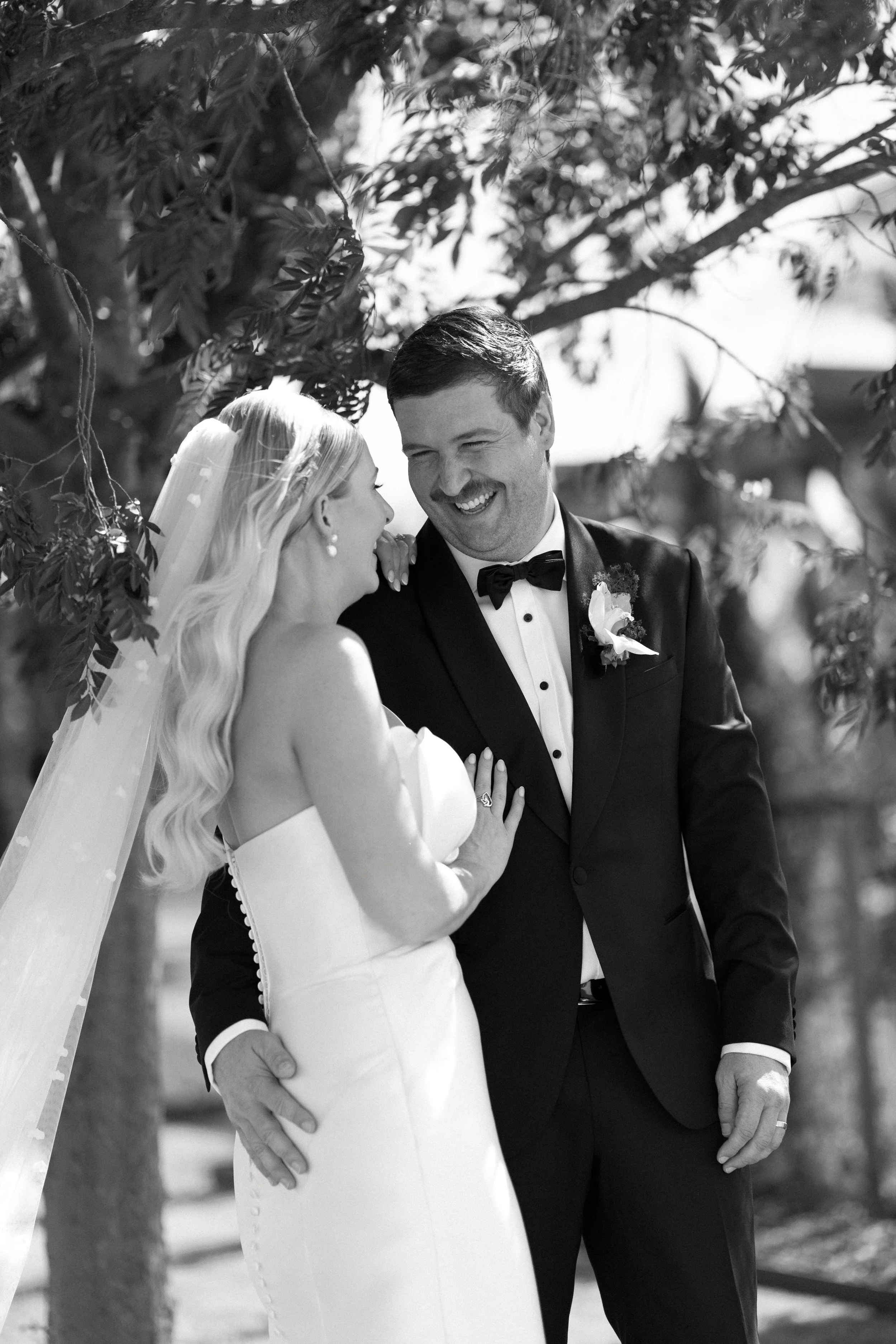 A black and white photo of a bride and groom smiling and looking at each other outdoors.