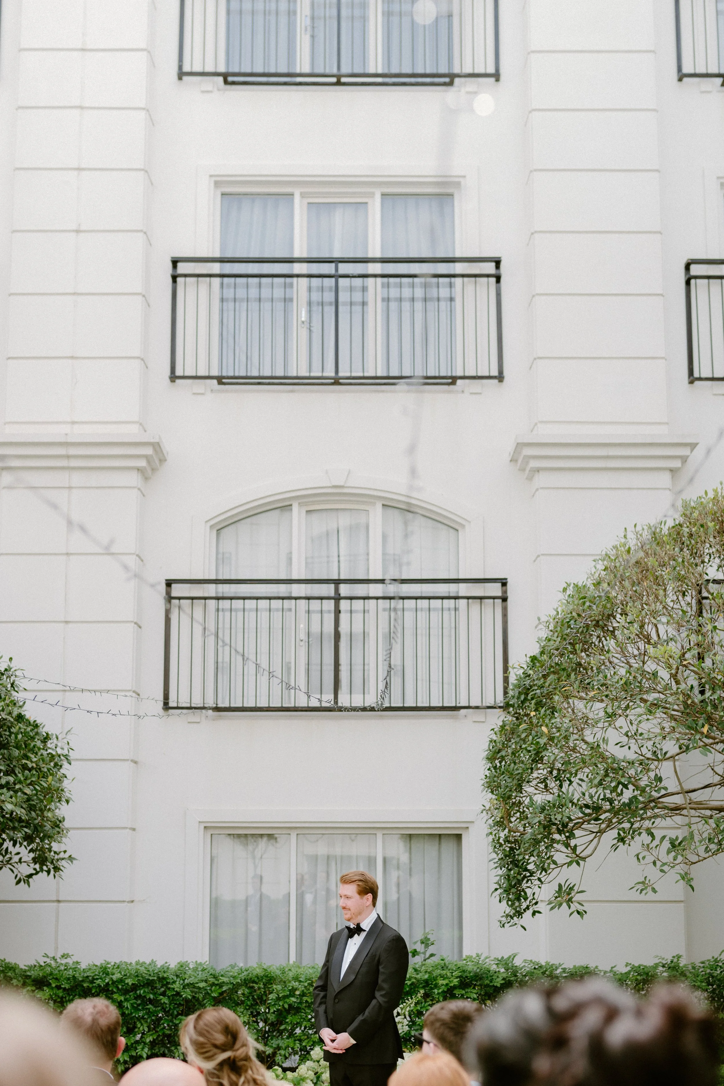 Man in a tuxedo standing outdoors in front of a group of people, in front of a white building with multiple balconies and large windows, with greenery around.