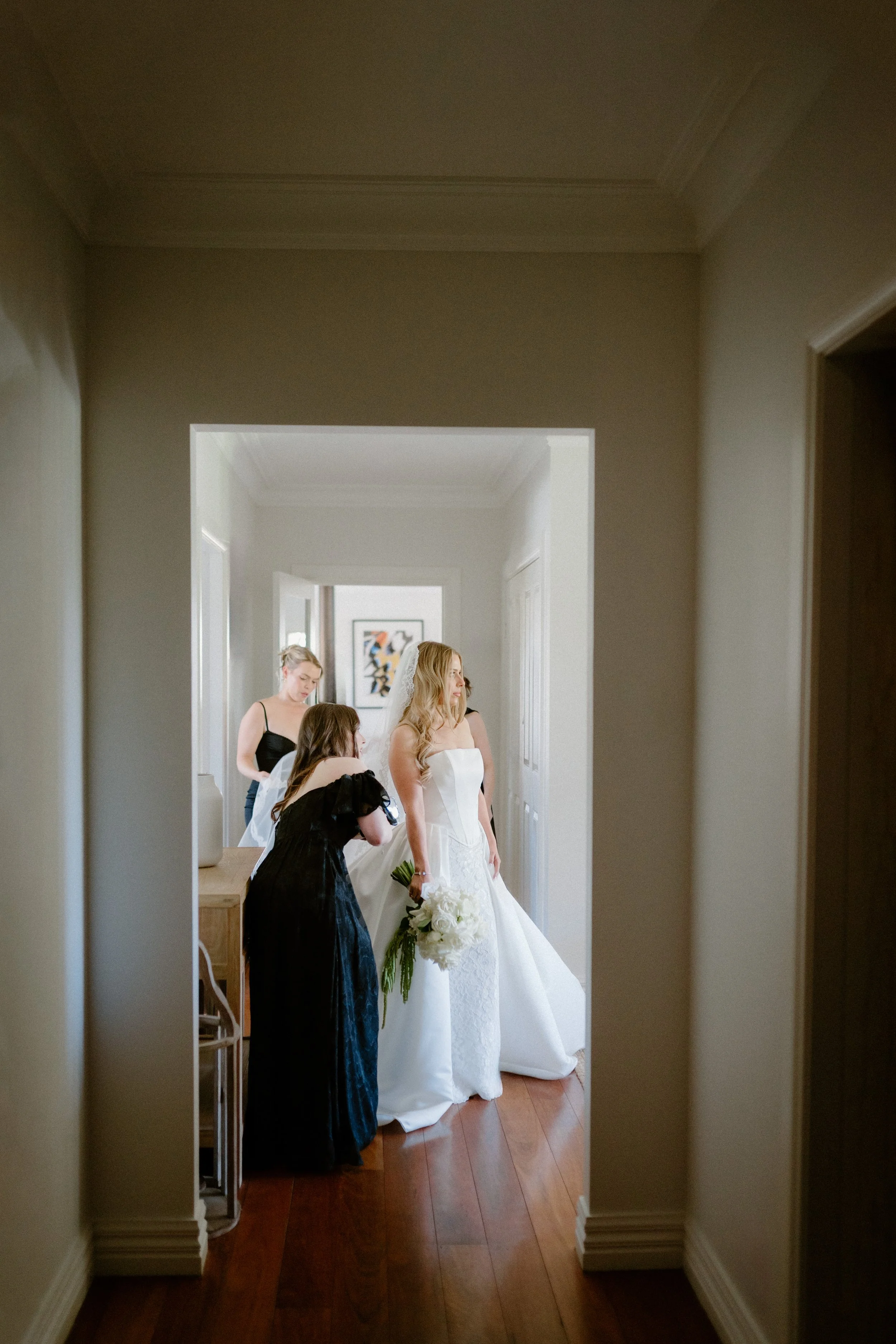 A bride in a white wedding dress holding a bouquet of white flowers, surrounded by her bridesmaids and friends, with one bridesmaid helping her adjust her dress, in a hallway of a house preparing for the wedding.