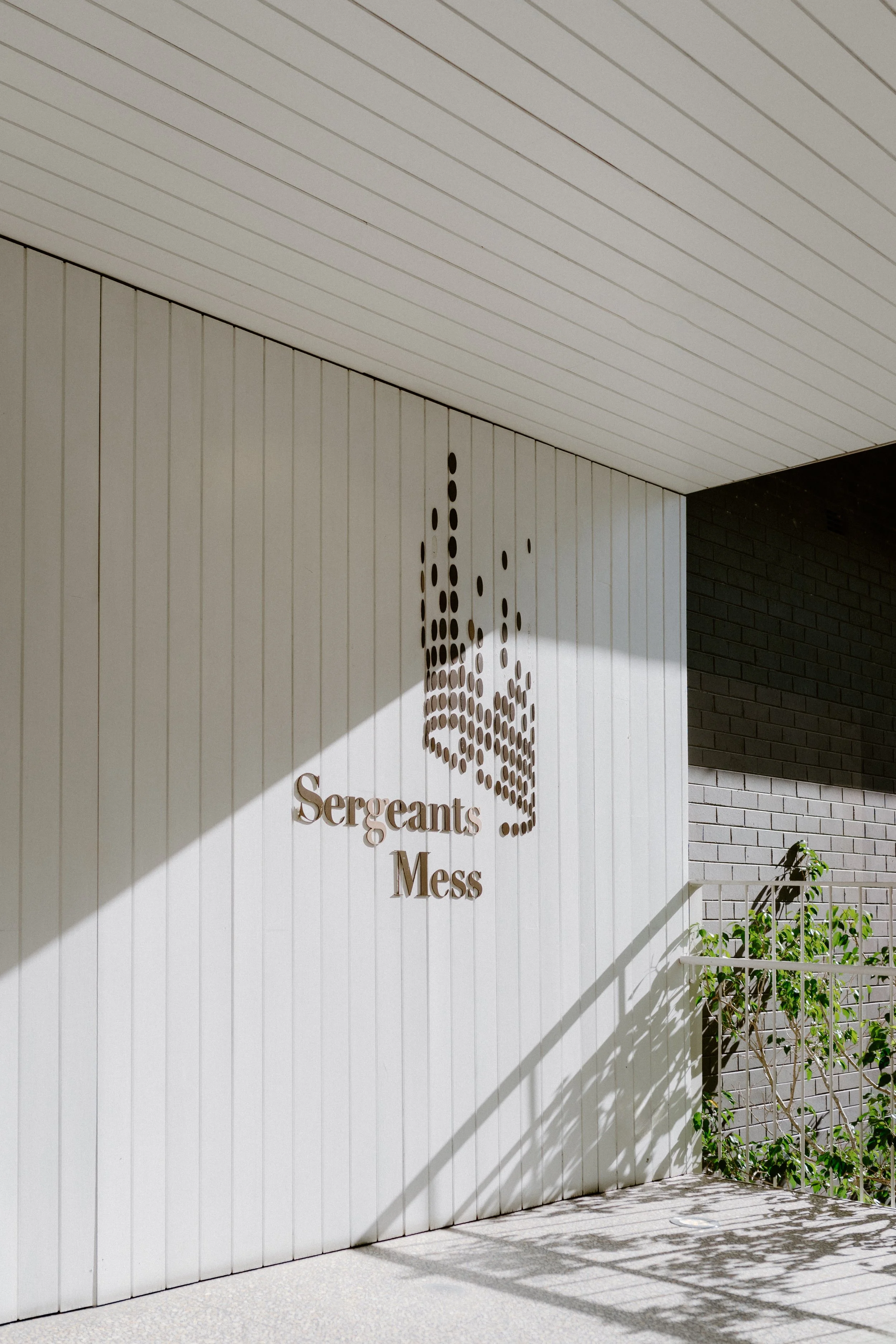 Exterior view of a building with a modern design, featuring a white vertical panel wall with a metal sign that says 'Sergeants Mess' and a stylized logo made of dots. Sunlight casts a shadow across the wall, and there are some green plants near the s