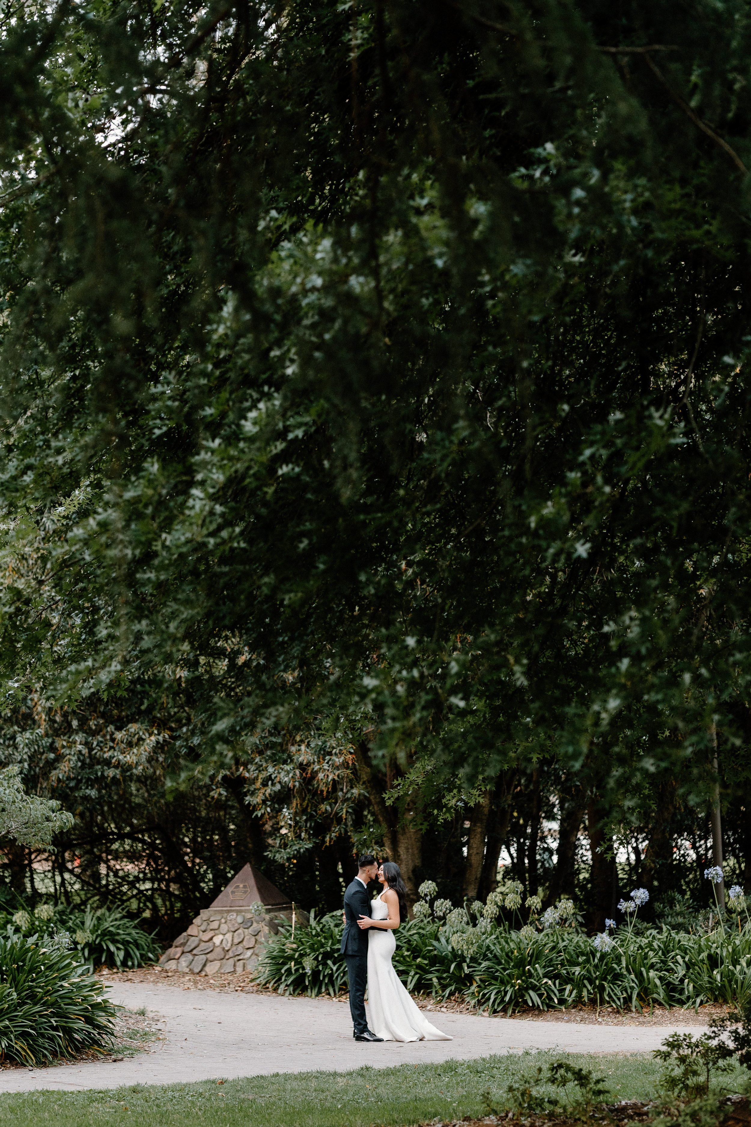 A bride and groom standing on a garden pathway, facing each other and holding hands, surrounded by lush greenery and flowers.