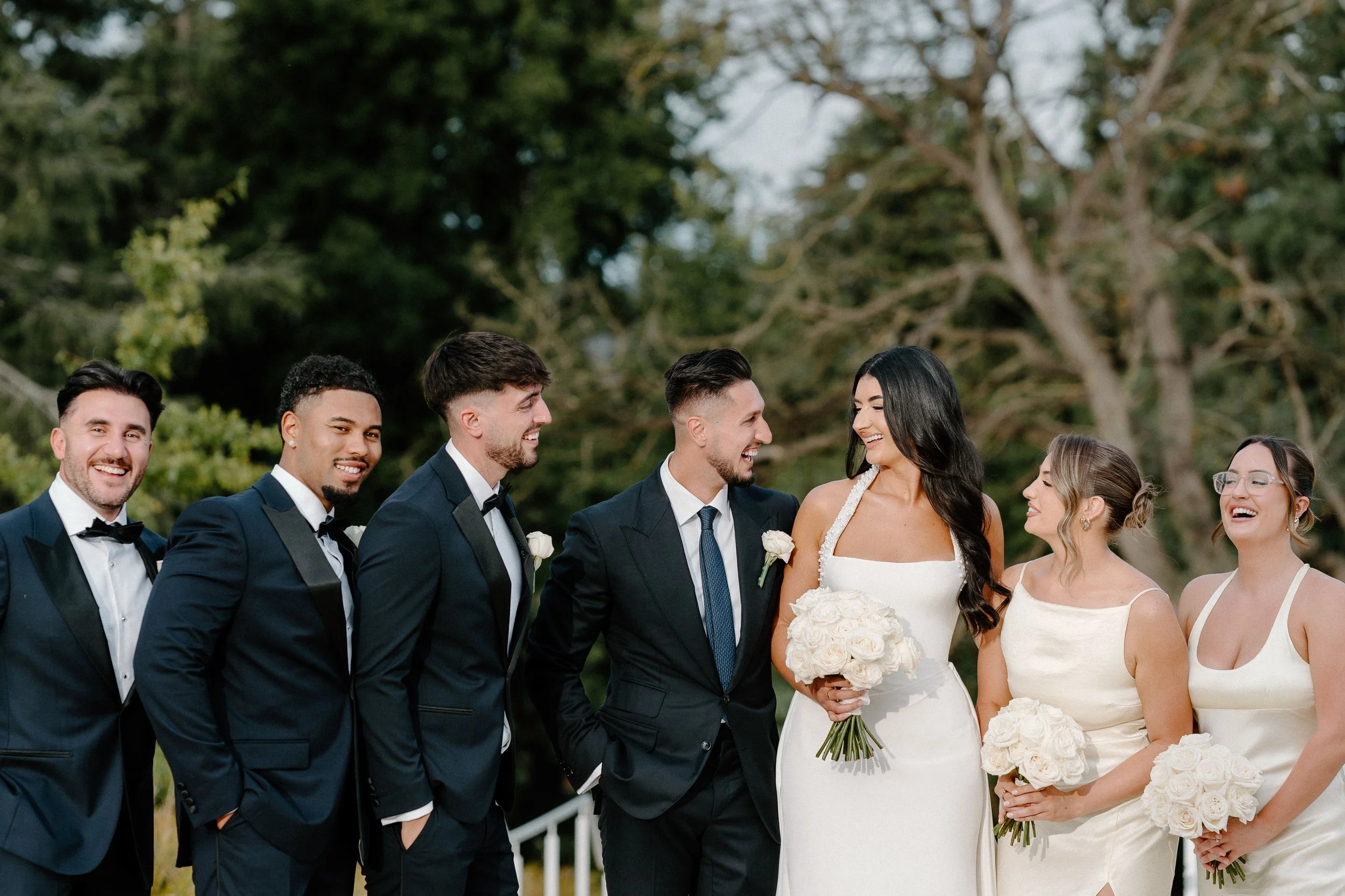 Wedding party: three groomsmen in black tuxedos with bow ties, and three bridesmaids in white dresses, standing outdoors with trees in the background, smiling and holding bouquets.