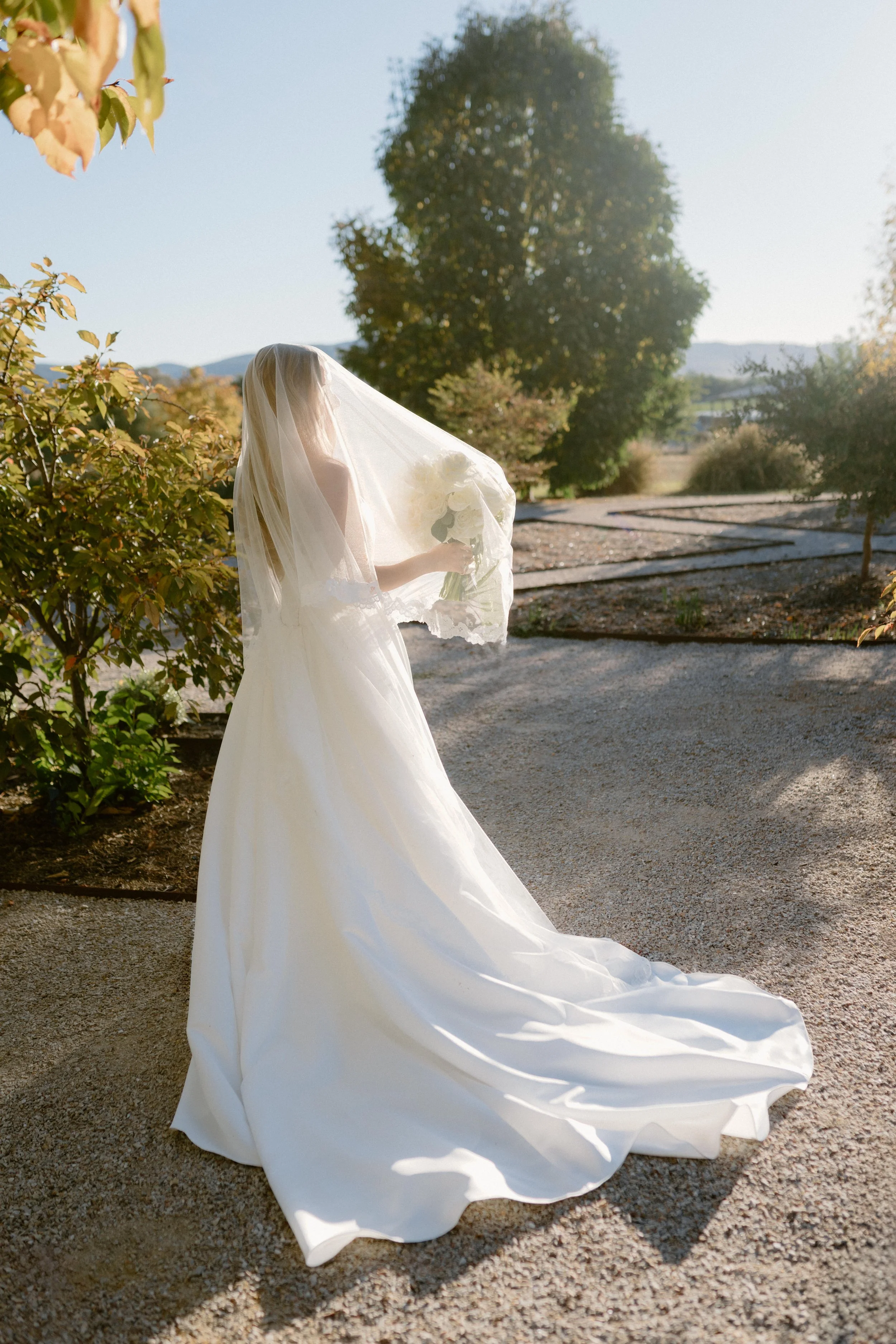A bride in a white wedding dress holding a bouquet of white flowers outdoors during daytime. The bride is under a sheer veil, and the dress has a long train on the ground. The background features trees and mountains, with clear sky and sunlight.