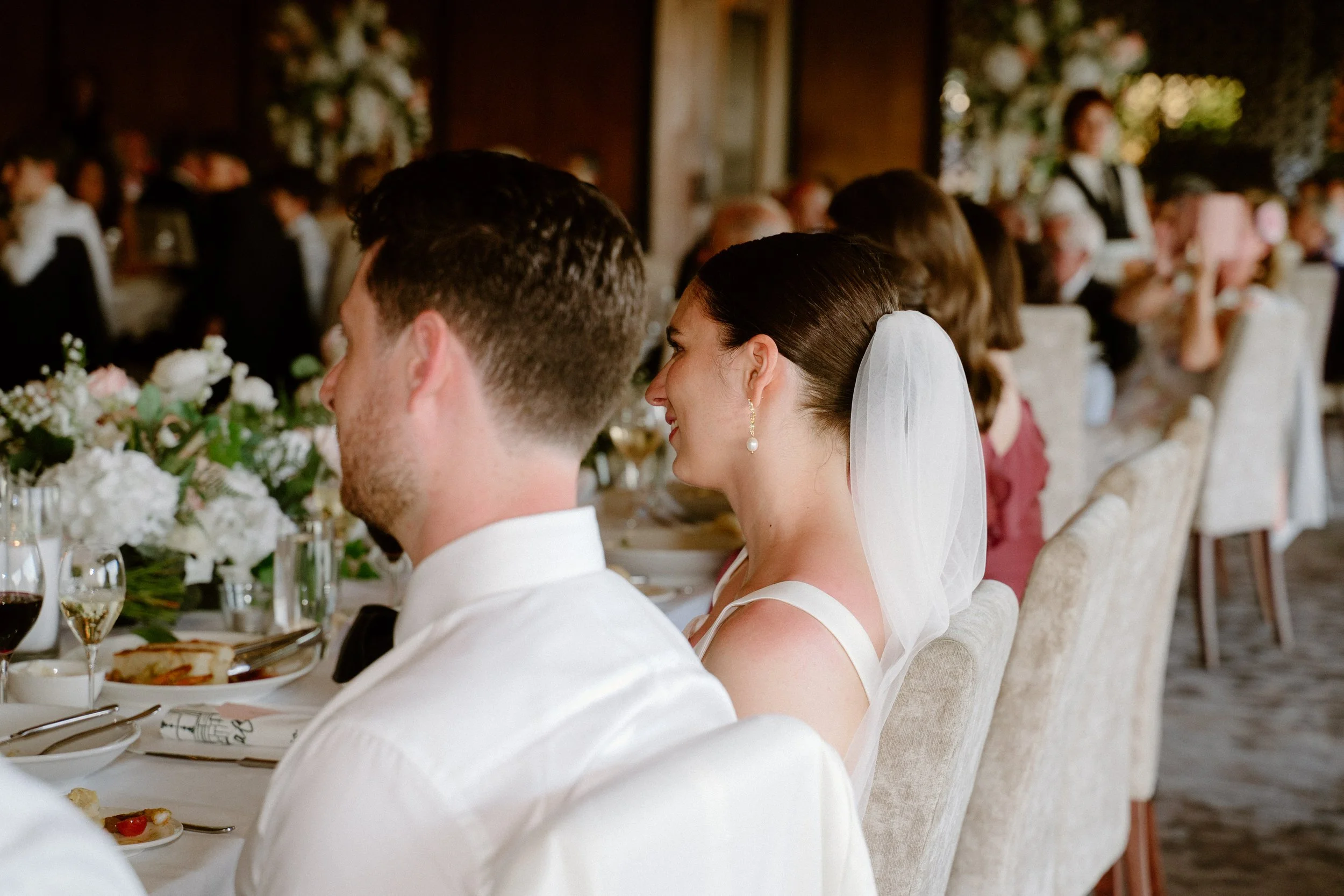 Bride and groom sitting at a wedding reception, smiling and listening to speeches, with guests seated at tables in the background.