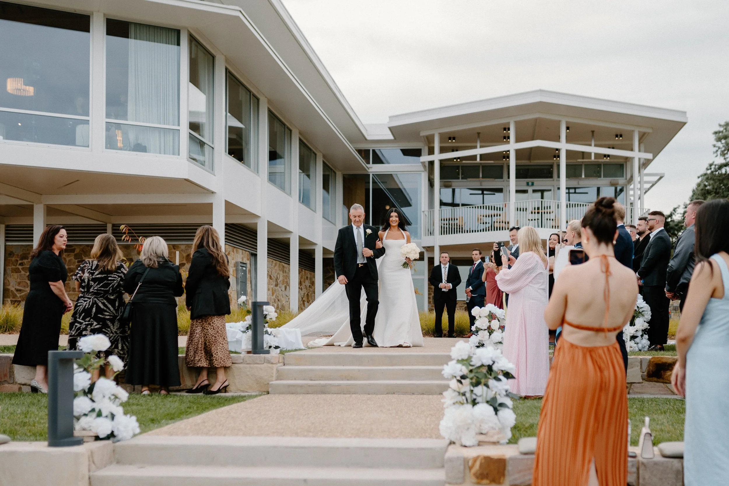 Bride walking down the stairs with her father at an outdoor wedding ceremony, surrounded by guests and floral decorations.
