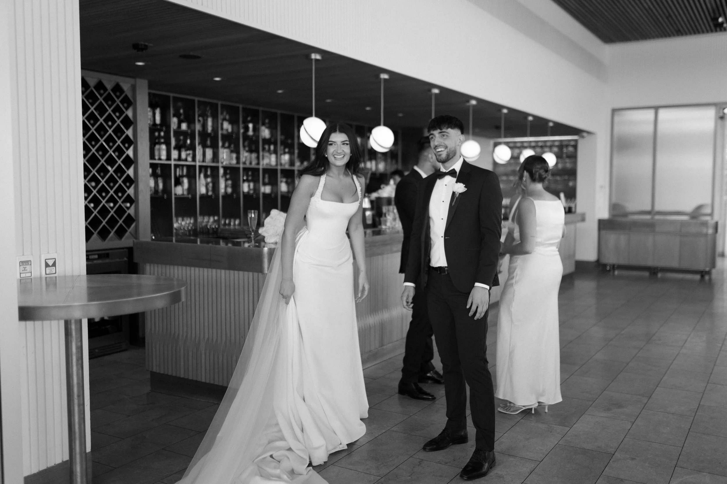 A bride and groom at their wedding reception, smiling and standing inside a modern venue with a bar and hanging lights, while two women are in the background.