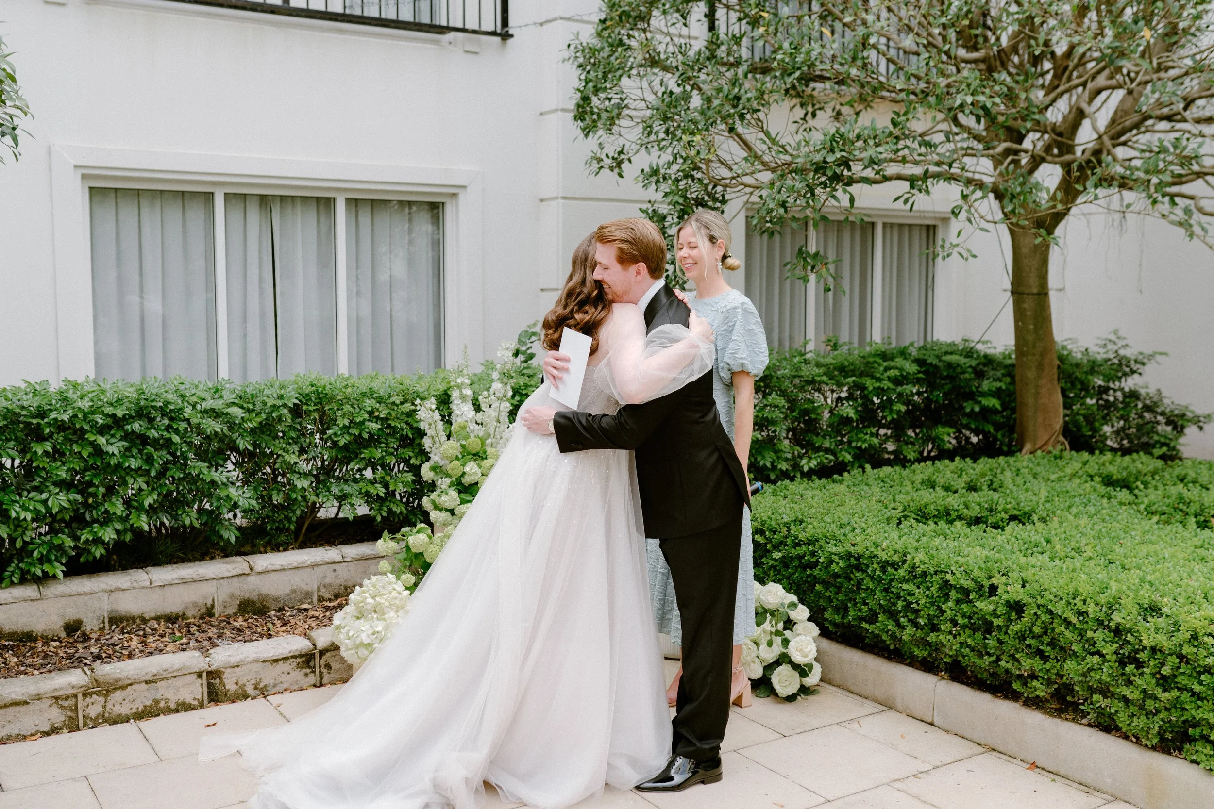 Bride and groom hugging during wedding ceremony in an outdoor garden, with an officiant and lush greenery in the background.