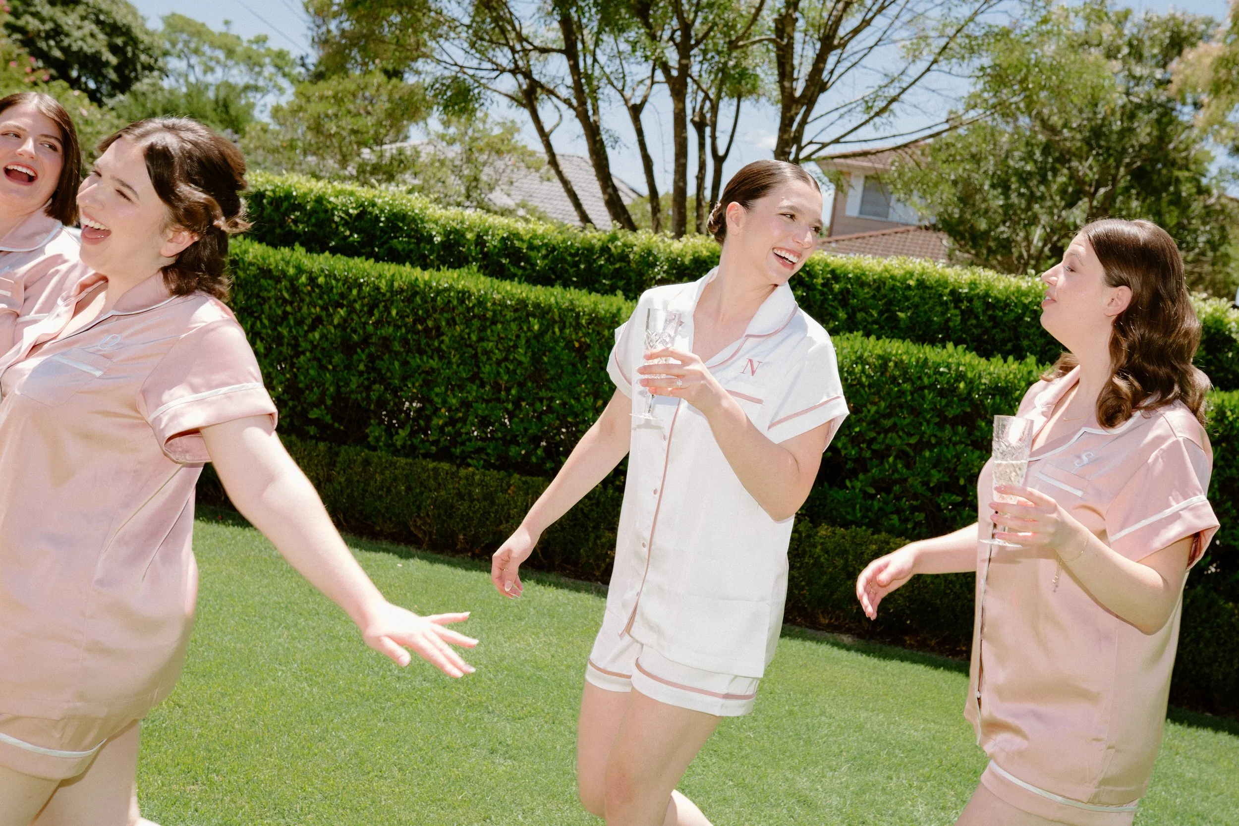 Group of women in pastel pajamas enjoying a sunny outdoor gathering, holding glasses of champagne and smiling.