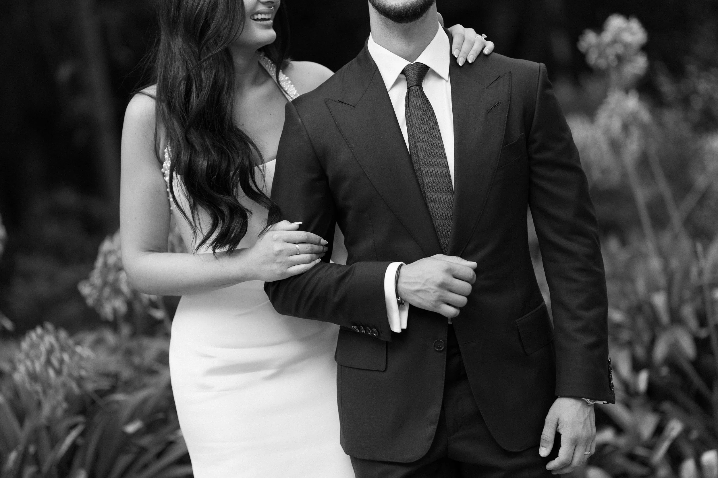 A black and white photo of a couple, with the woman smiling and her arm on the man's shoulder, dressed in wedding attire outdoors.