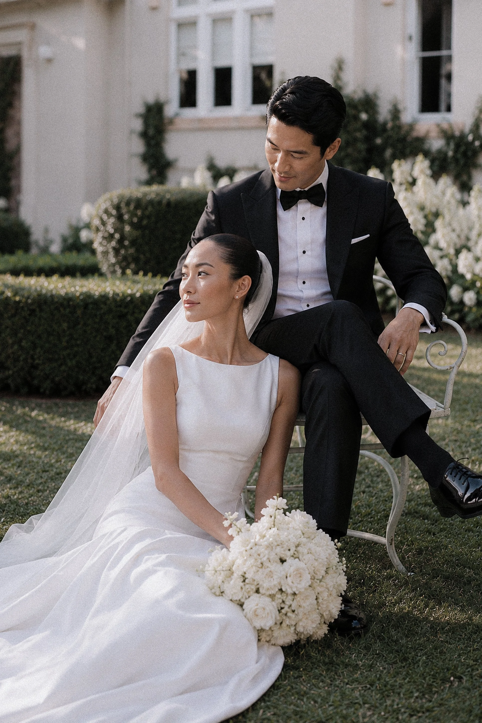 A bride and groom sitting outdoors on a white bench, with the bride holding a bouquet of white flowers. The bride is wearing a white wedding gown with a veil, and the groom is dressed in a black tuxedo with a bowtie. They are in front of a white hous