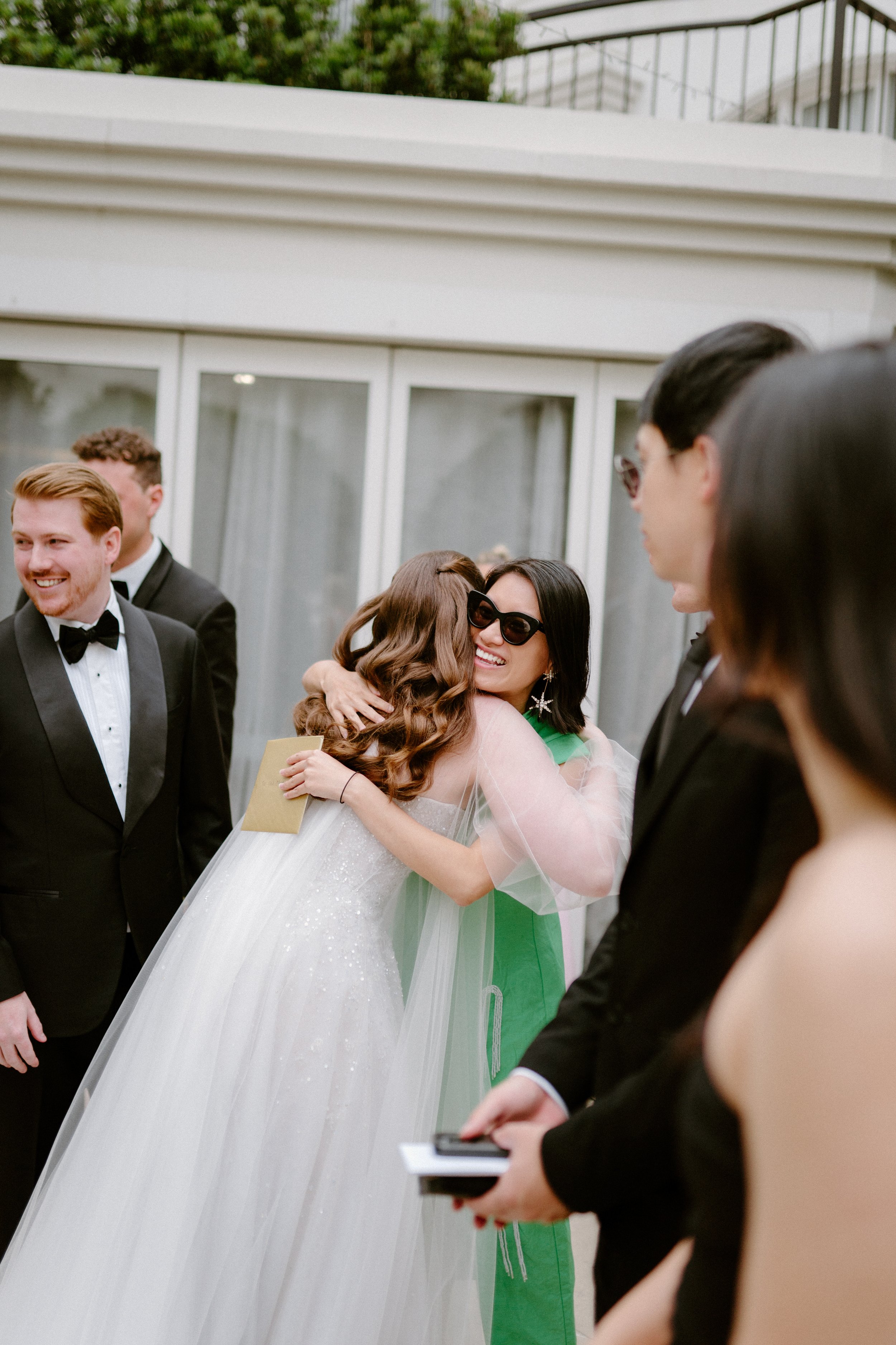 Two women hugging at a wedding reception, one in a white wedding gown and the other in a green dress, surrounded by men in tuxedos.