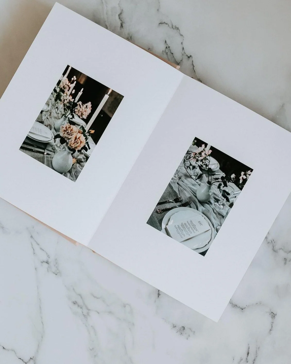 Open the white photo album showing two pages, each with a photo of a table setting decorated with flowers and candlesticks, on a marble surface.