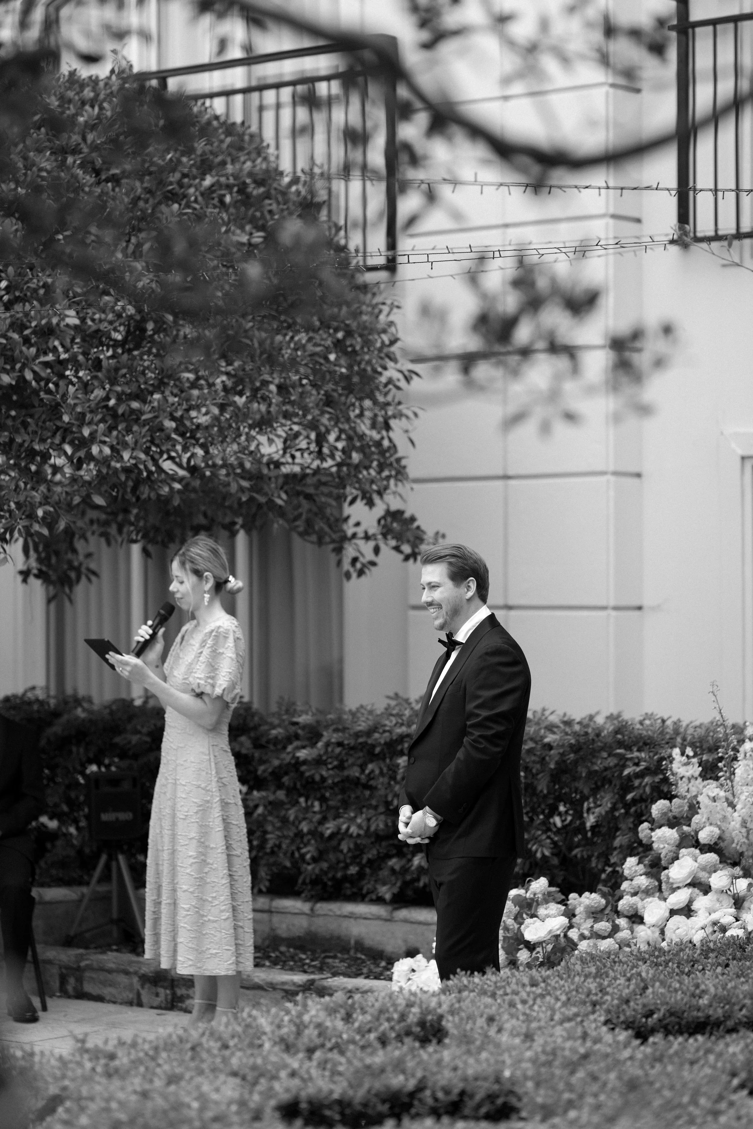 A black and white photo of a wedding ceremony outdoors, with a woman reading vows on a microphone and a smiling groom in a tuxedo standing nearby.