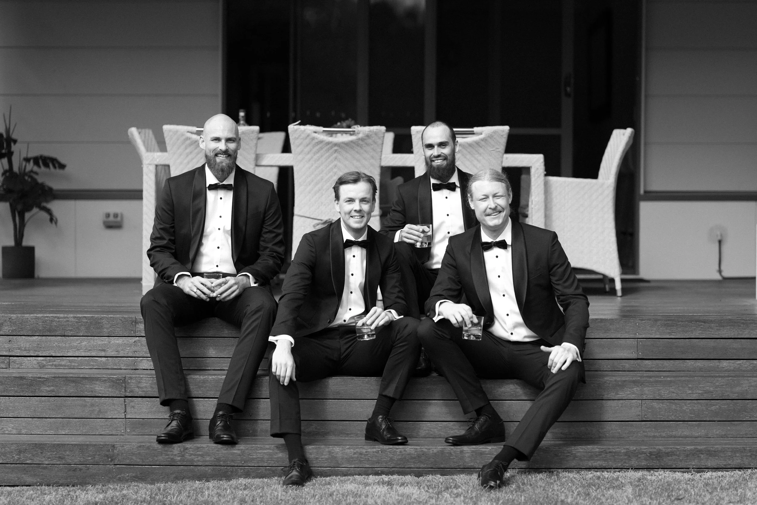 Black and white photo of five men in tuxedos, some holding drinks, smiling and sitting on wooden steps in front of a dining area with chairs.