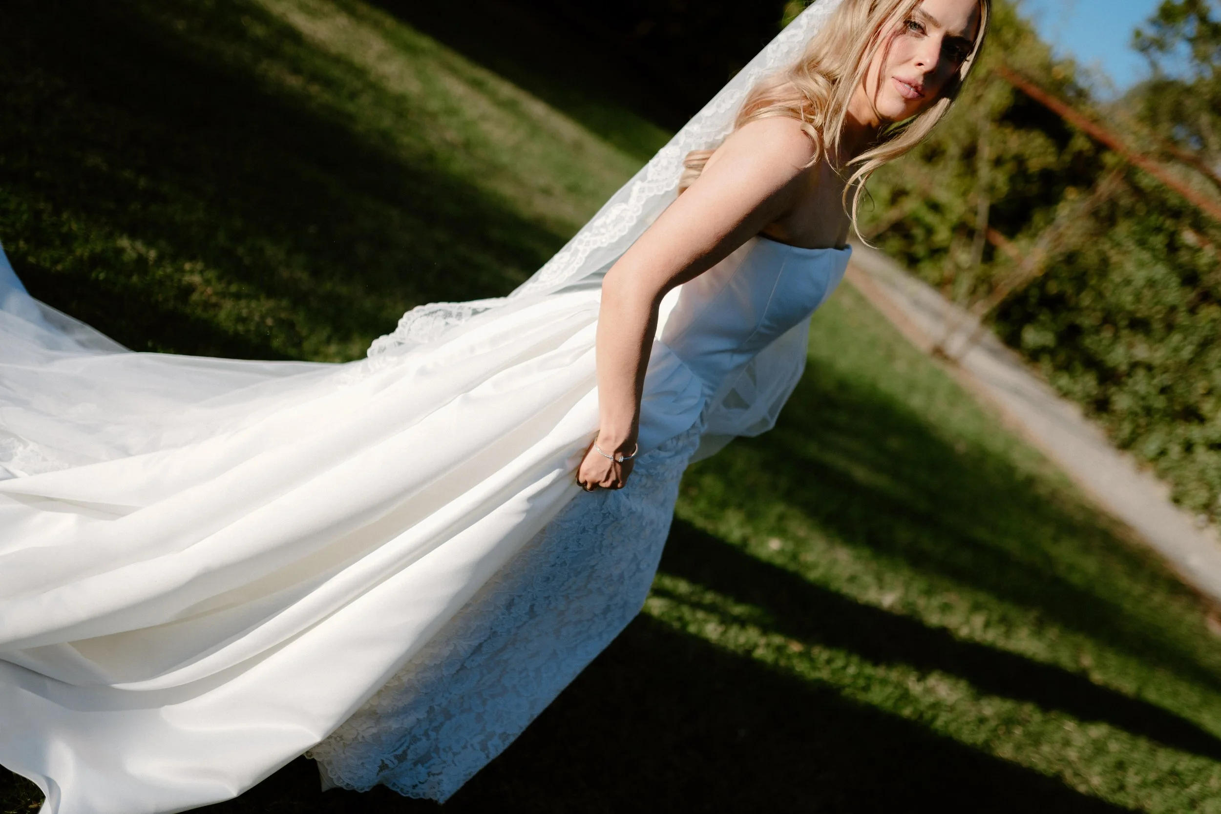 A woman in a white wedding dress outdoors in a grassy area under blue sky with trees and a building in the background.