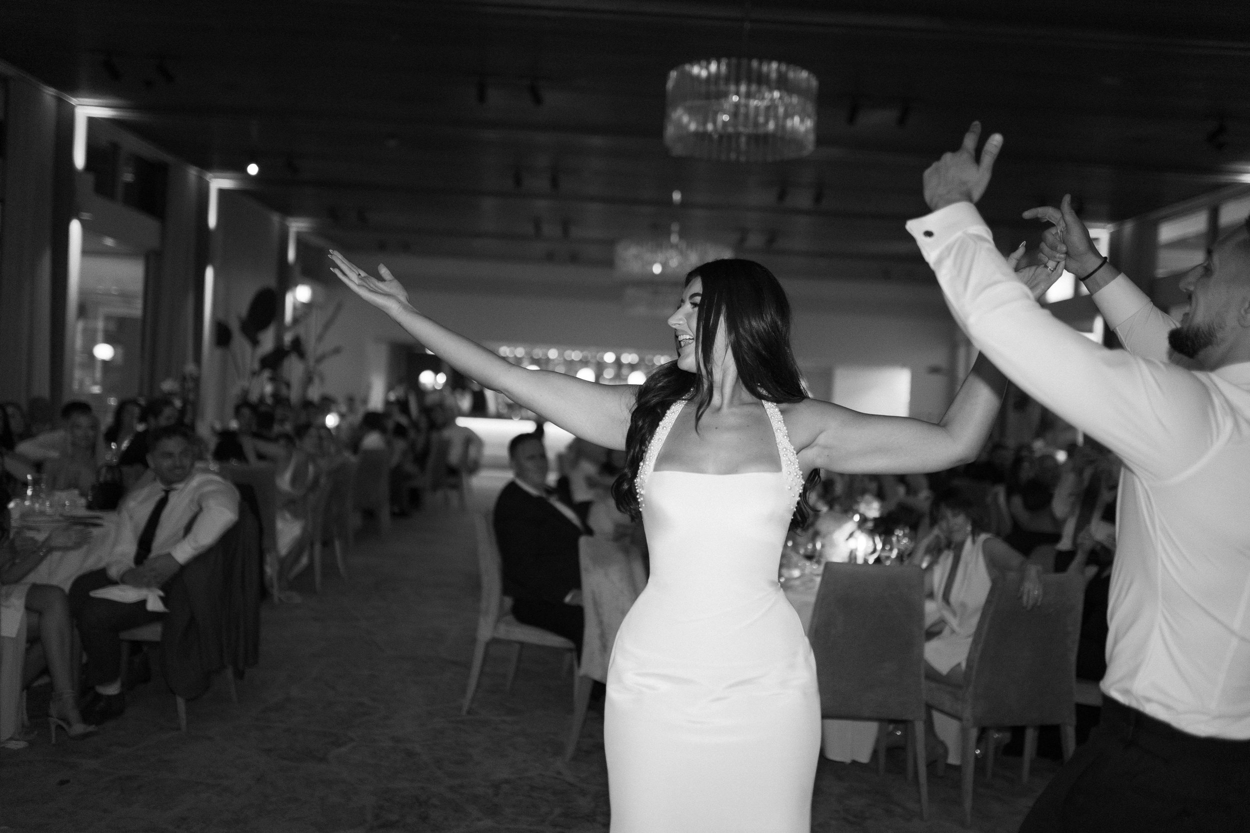 Black and white photo of a woman in a sleeveless wedding dress dancing at a wedding reception with guests seated at tables around her.