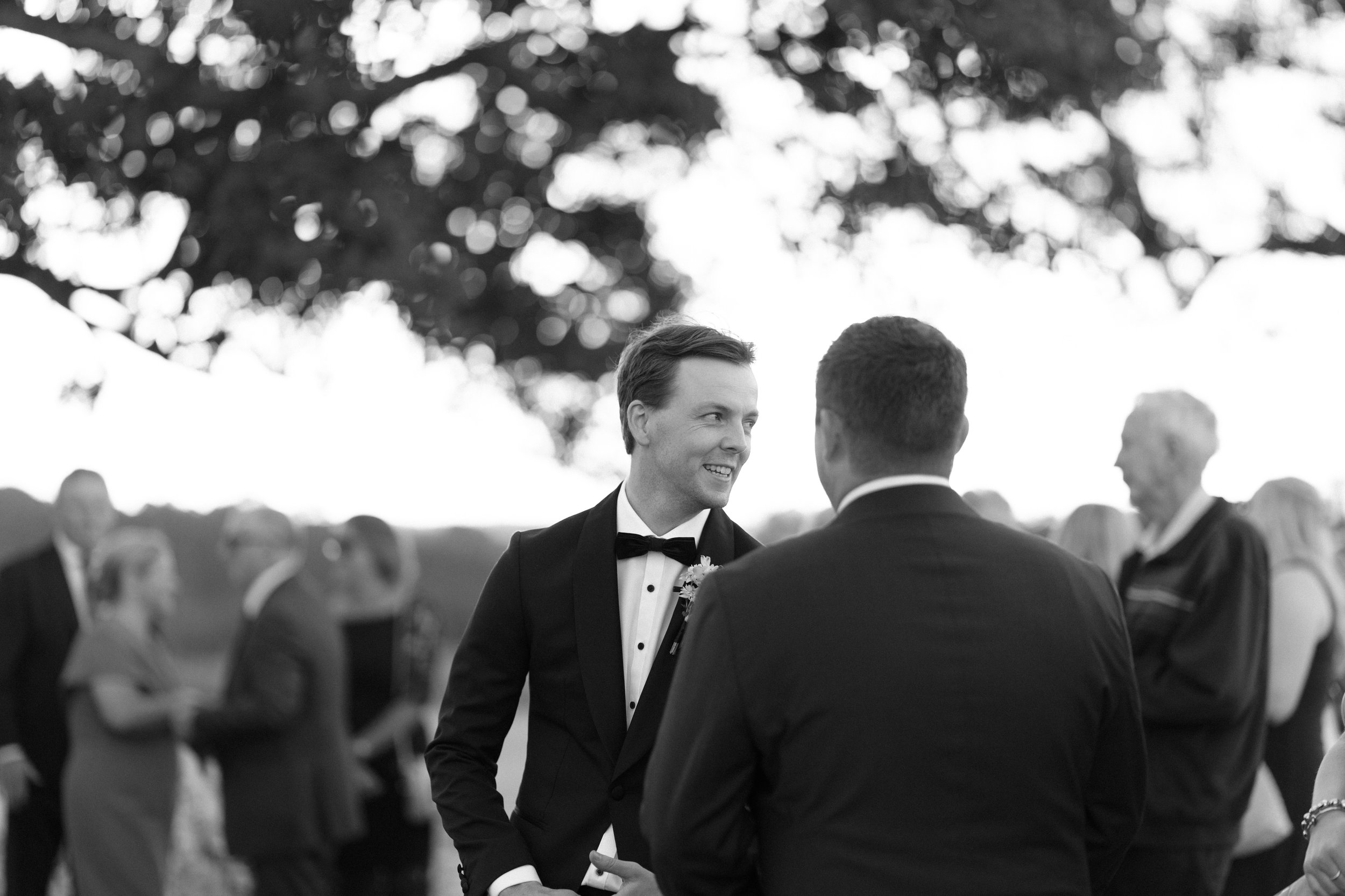 A black and white photograph of a wedding ceremony outdoors. The image focuses on a smiling man in a tuxedo, talking to another man. People are gathered in the background, some smiling and mingling, with trees and dappled sunlight overhead.